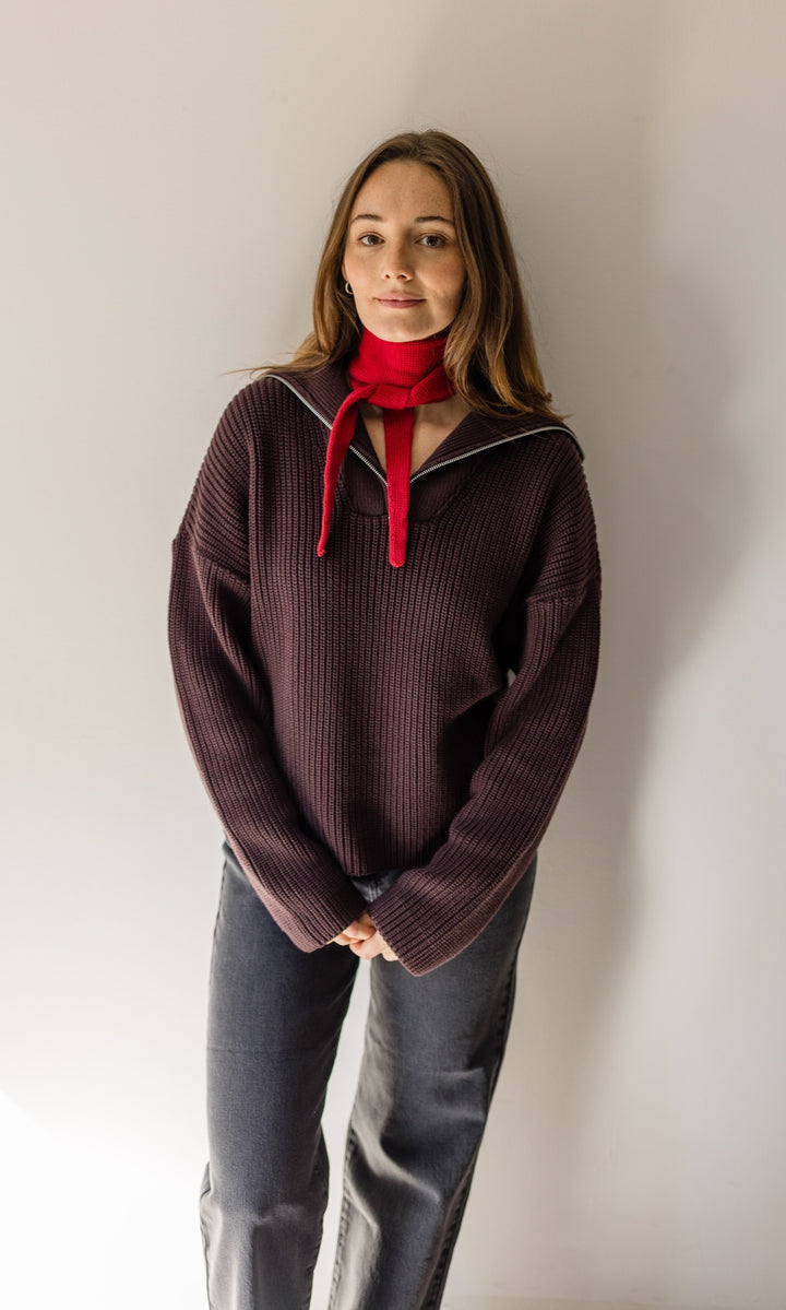 Gerippter Pullover mit Zipper Reissverschluss in braun mit dunkler Jeans und rotem kleinen Strickschal vor heller Wand Blick gerade