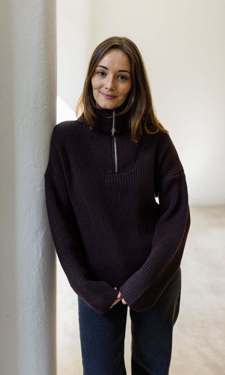 Gerippter Pullover mit Zipper Reissverschluss in braun mit dunkler Jeans vor heller Wand Blick lächelnd