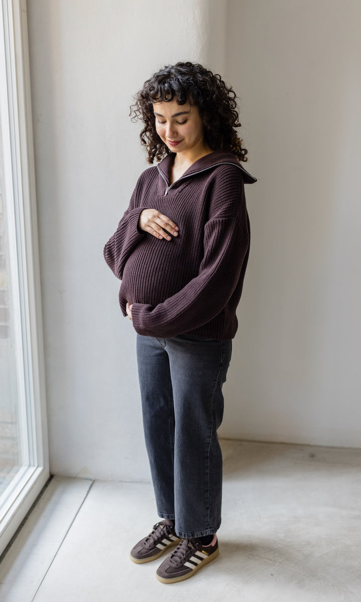Gerippter Pulli mit Zipper Reissverschluss in braun mit schwarzer Jeans und schwarzen Stiefeln schwanger vor heller Wand Blick auf Bauch