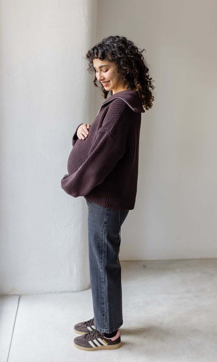 Gerippter Pulli mit Zipper Reissverschluss in braun mit schwarzer Jeans und braunen Sneakers schwanger seitlich vor heller Wand Blick auf Bauch lächelnd