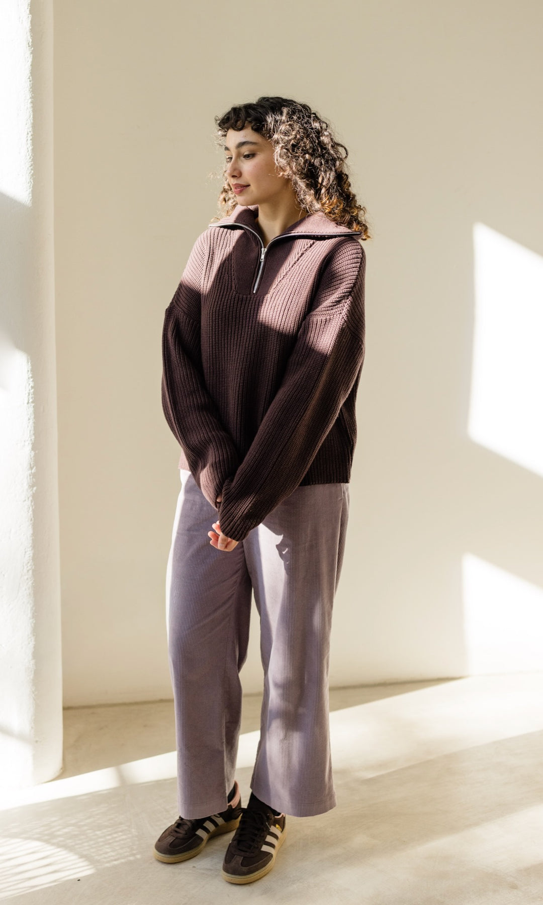 Gerippter Pullover mit Zipper Reissverschluss in braun mit hellbrauner Hose und braunen Sneakers vor heller Wand seitlich
