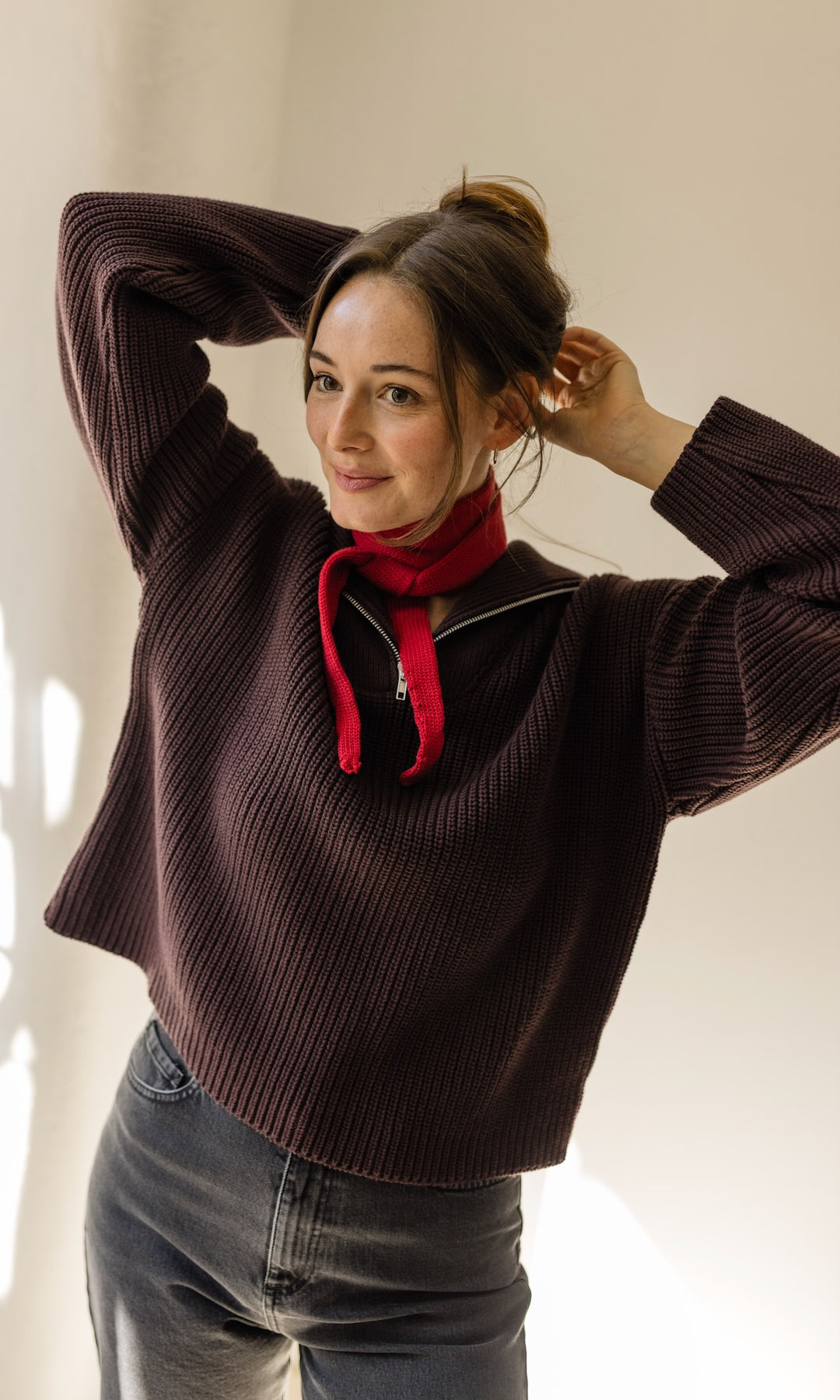 Gerippter Pullover mit Zipper Reissverschluss in braun mit dunkler Jeans und rotem kleinen Strickschal vor heller Wand Blick seitlich