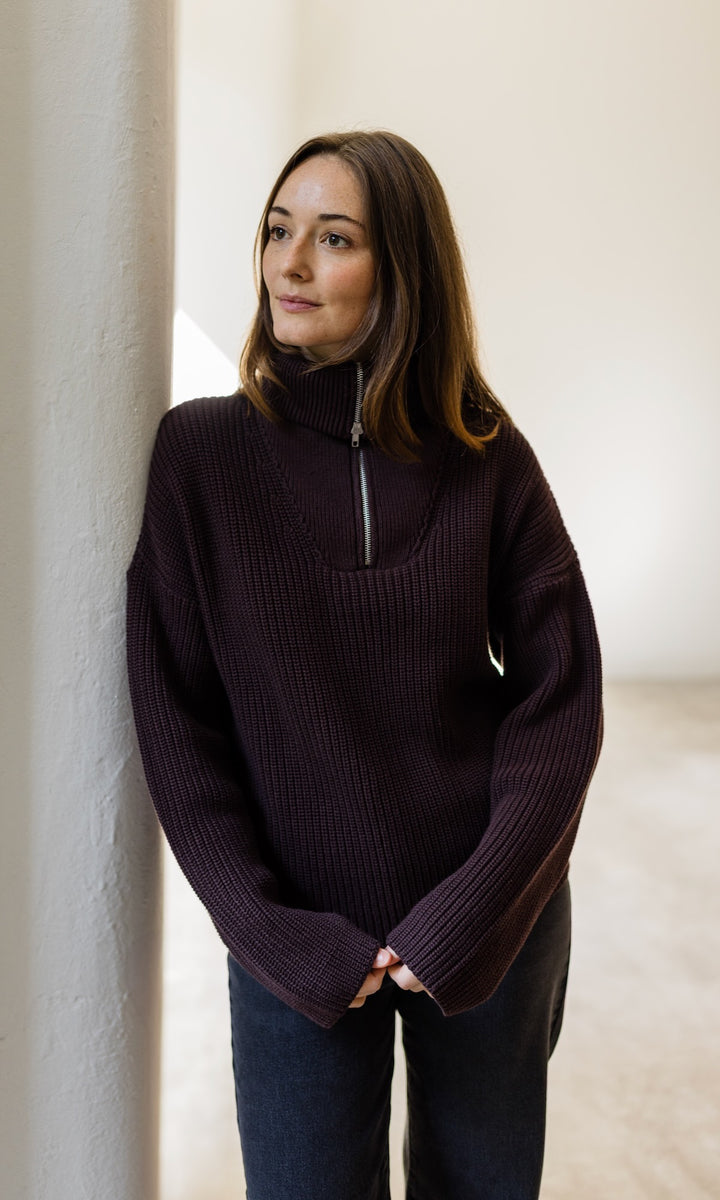 Gerippter Pullover mit Zipper Reissverschluss in braun mit dunkler Jeans vor heller Wand Blick nach oben