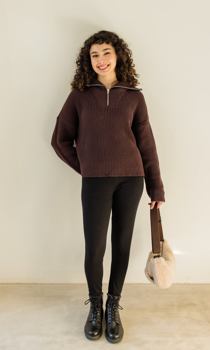 Gerippter Pulli mit Zipper Reissverschluss in braun mit schwarzer Leggings und braunen Stiefeln vor heller Wand Blick gerade lachend