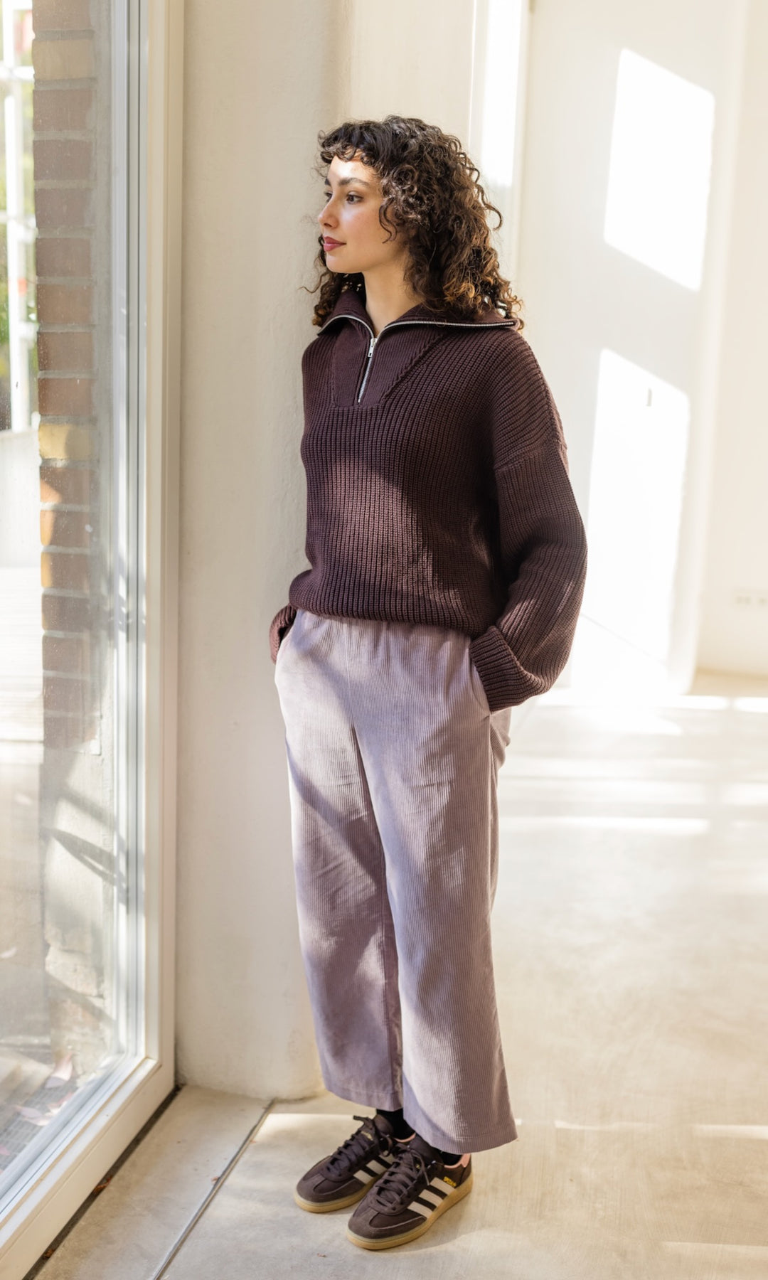 Gerippter Pullover mit Zipper Reissverschluss in braun mit hellbrauner Hose und braunen Sneakers vor heller Wand Blick aus Fenster