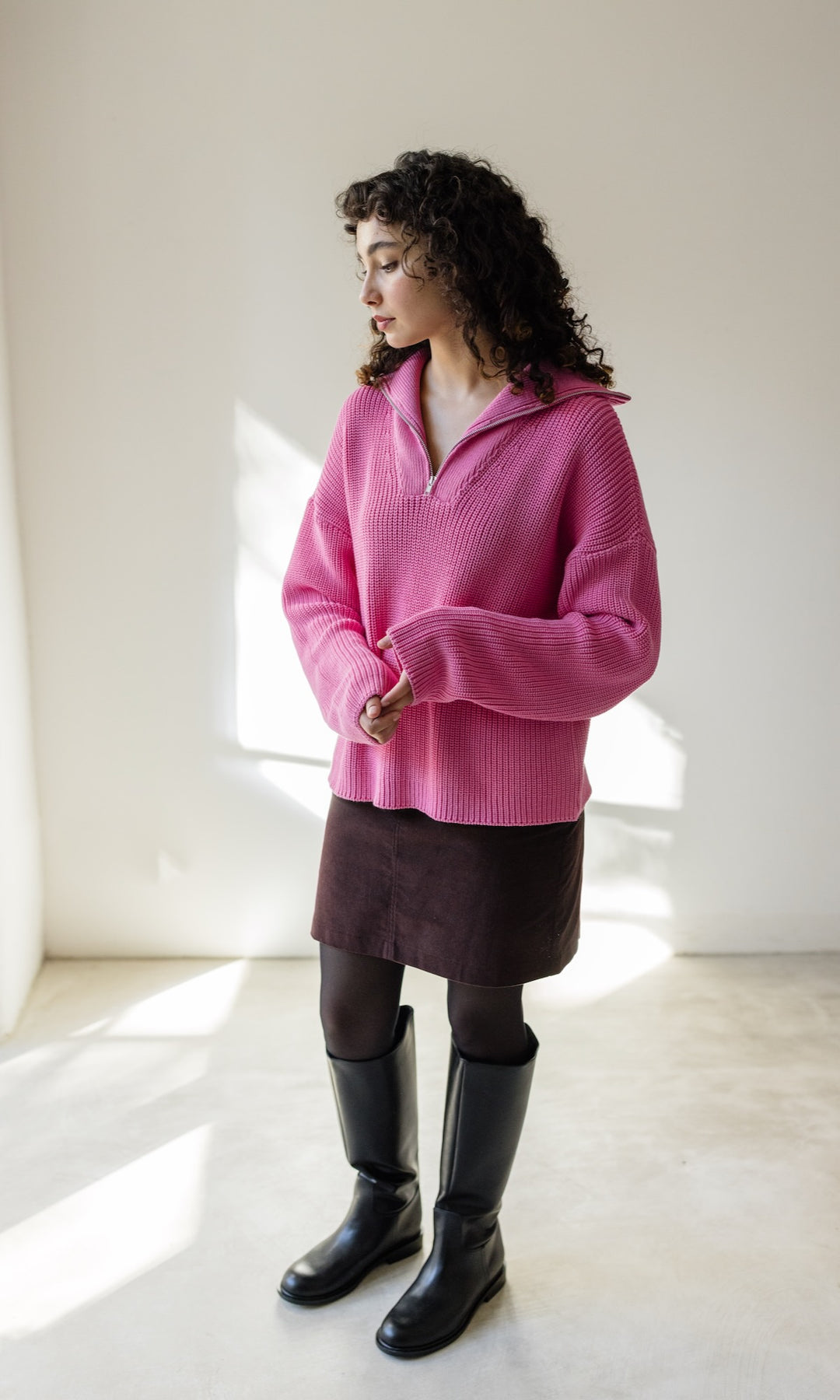 Gerippter Pullover mit Zipper Reissverschluss in rosa mit braunem Rock und schwarzen Stiefeln vor heller Wand Blick seitlich nach unten