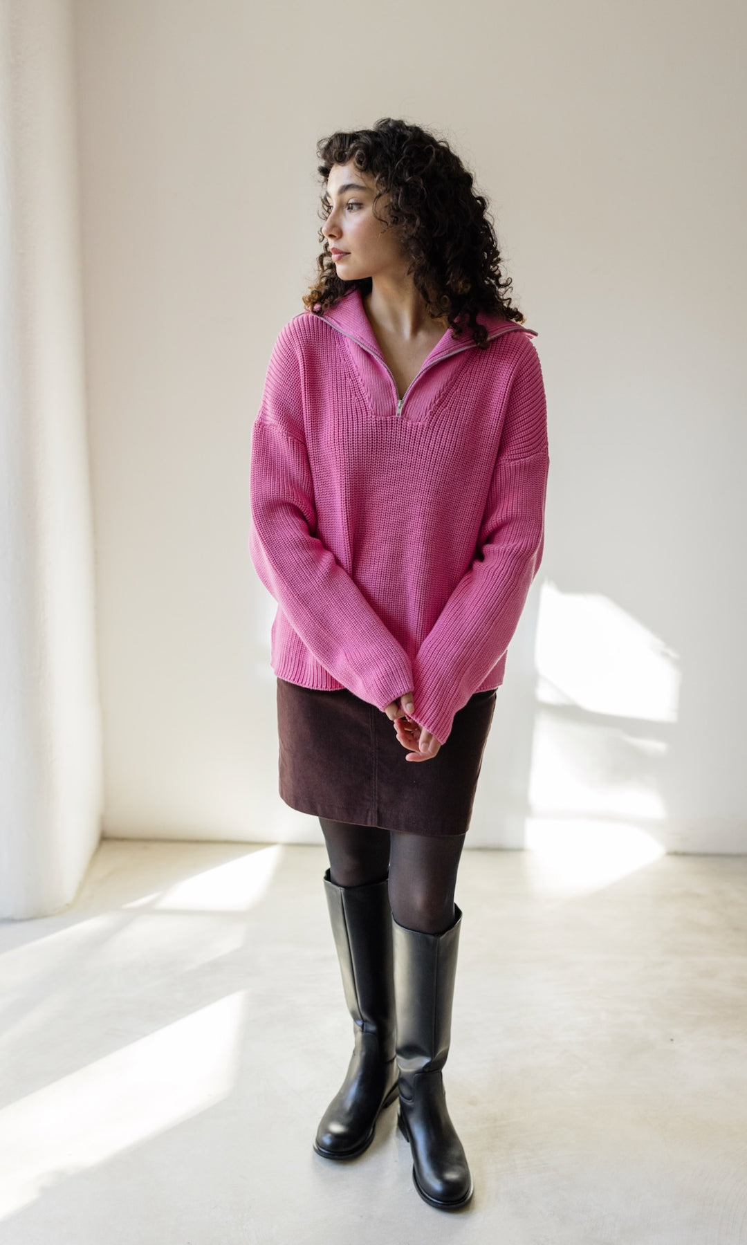 Gerippter Pullover mit Zipper Reissverschluss in rosa mit braunem Rock und schwarzen Stiefeln vor heller Wand Blick seitlichaus Fenster