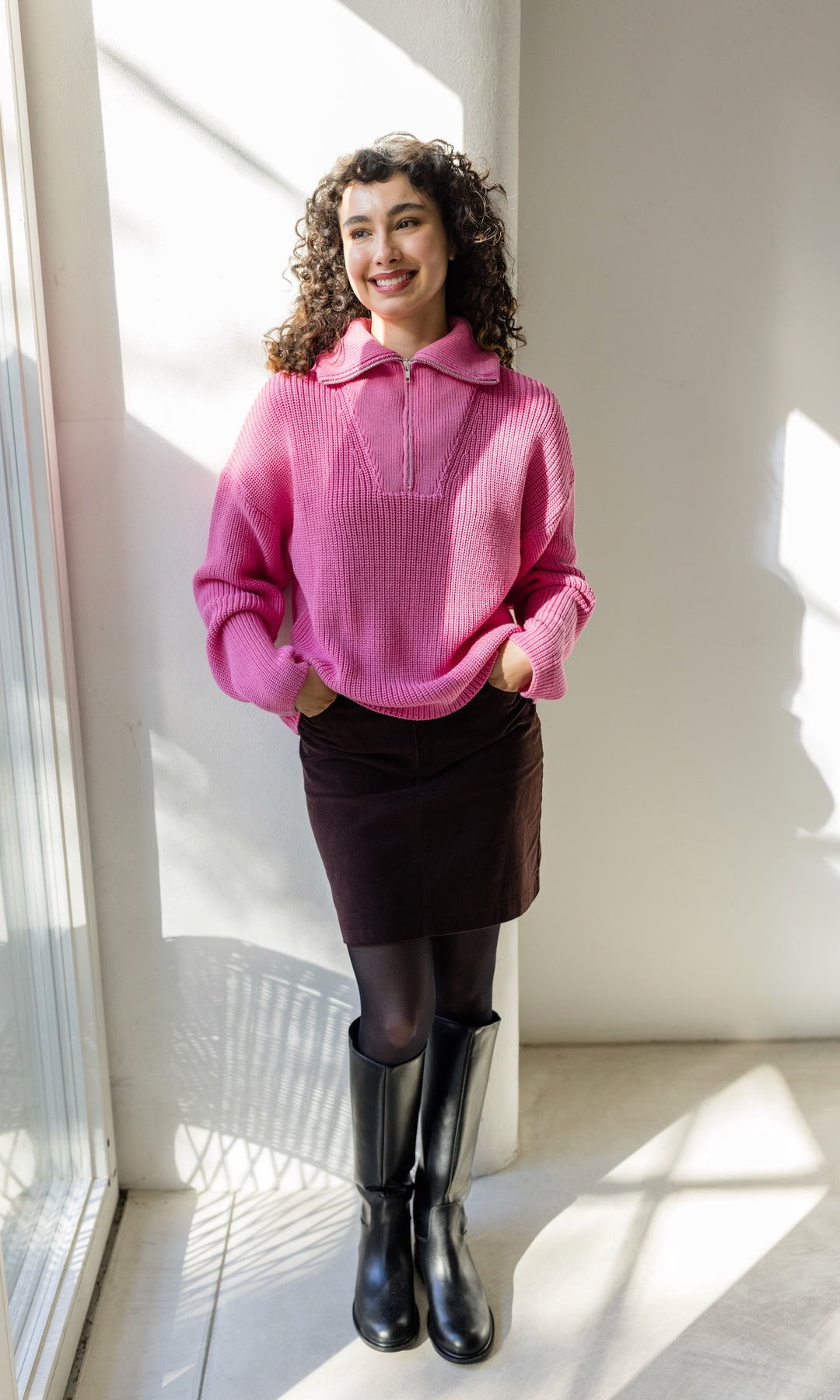 Gerippter Pullover mit Zipper Reissverschluss in rosa mit braunem Rock vor heller Wand Blick lächelnd aus Fenster