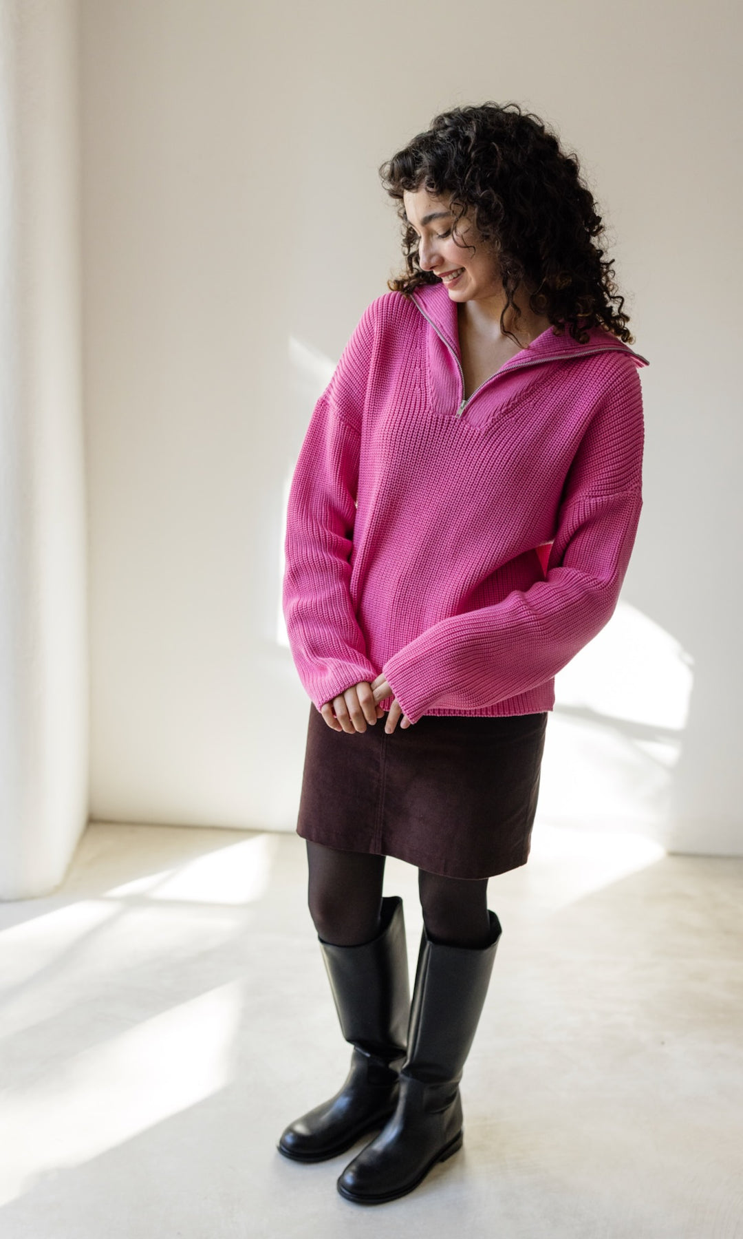 Gerippter Pullover mit Zipper Reissverschluss in rosa mit braunem kleinem Strickschal und braunem Rock und schwarzen Stiefeln vor heller Wand Blick nach unten