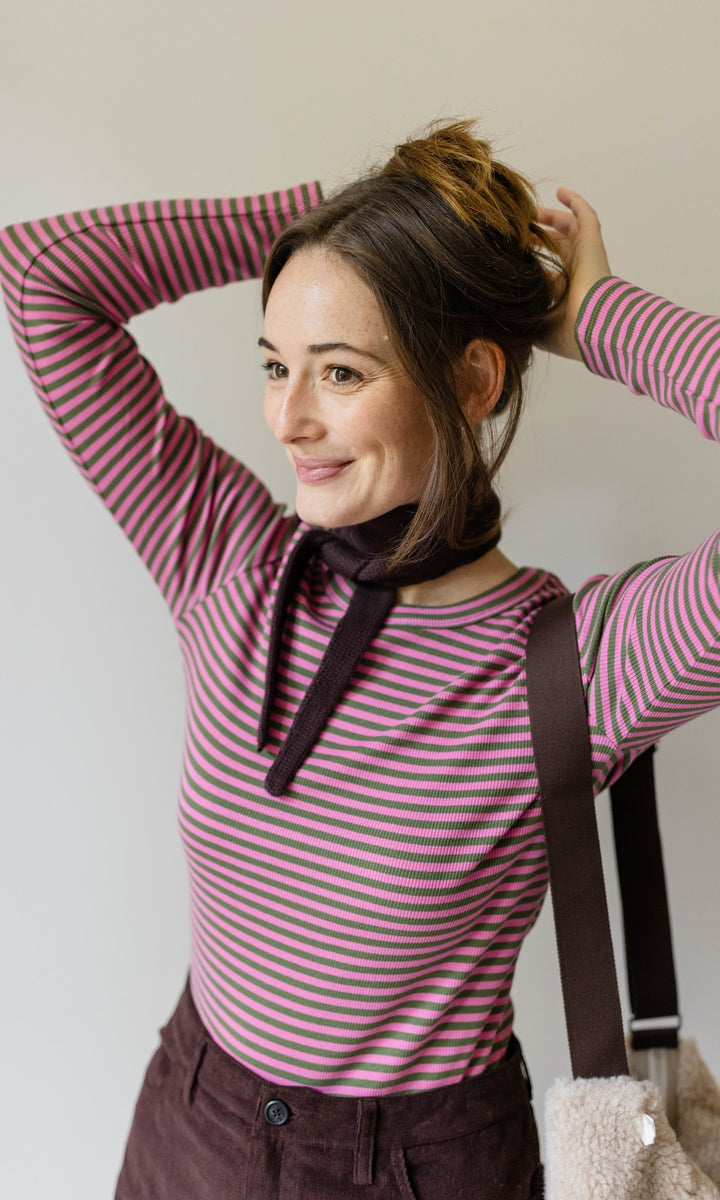 Kleiner Mini Strickschal in braun im Outfit mit rosa gestreiftem Shirt, braunem Rock und Woll Handtasche vor heller Wand Blick seitlich lächelnd