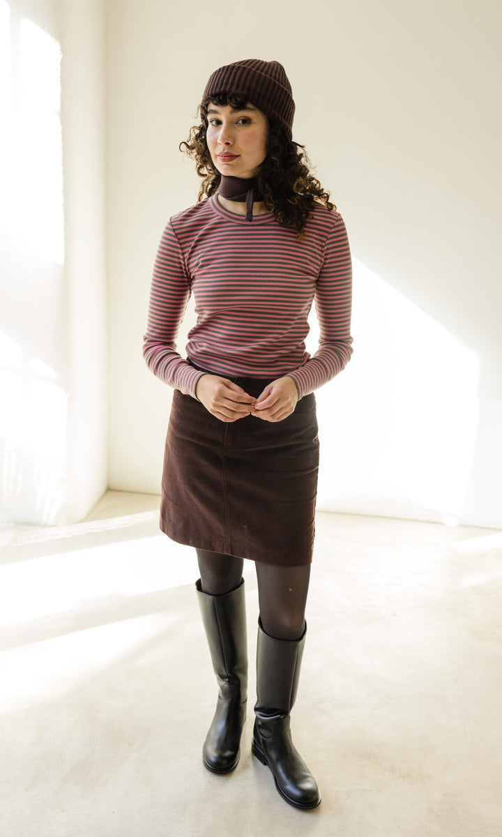 Kleiner Mini Strickschal in braun im Outfit mit rosa gestreiftem Shirt, braunem Rock und schwarzen Stiefeln vor heller Wand Blick gerade