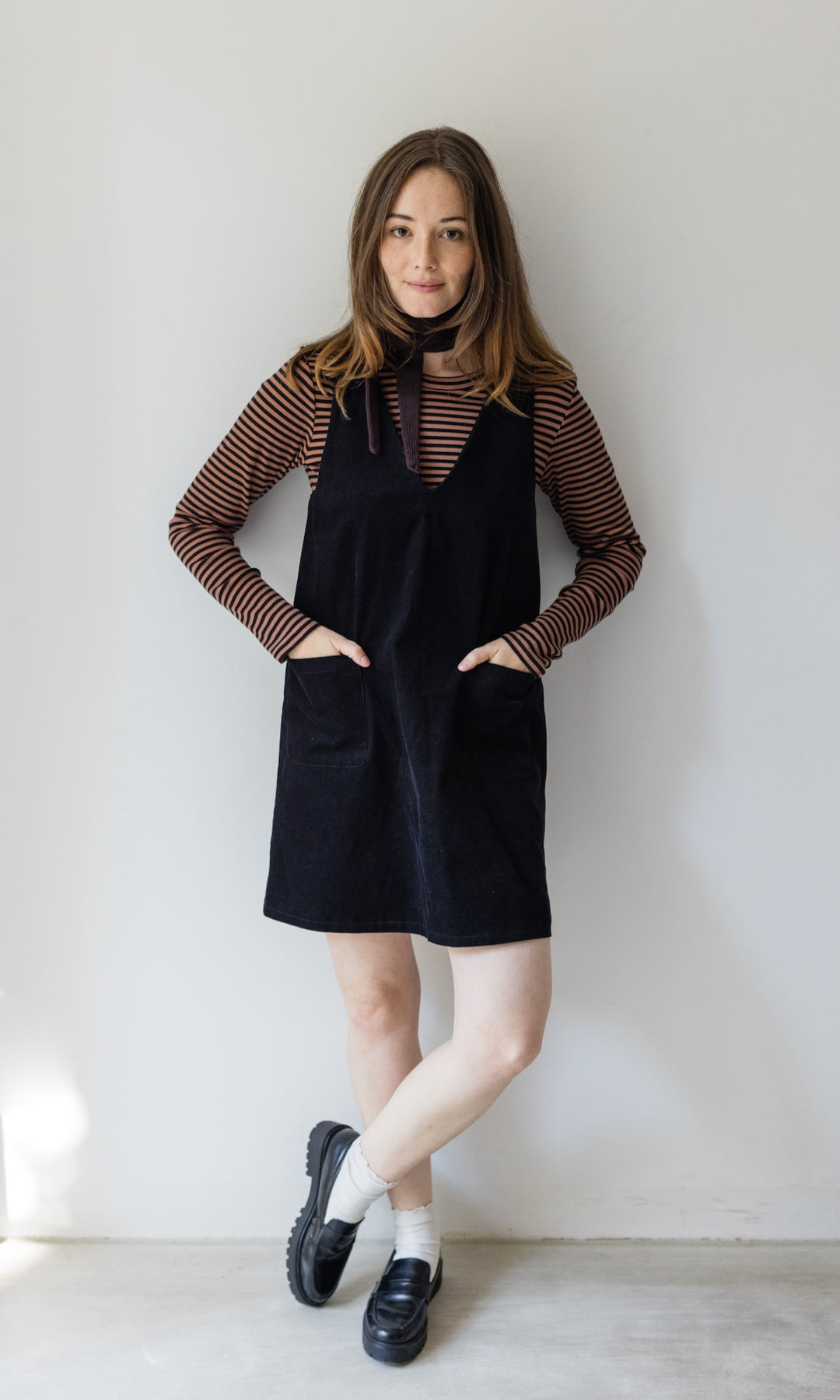 Kleiner Mini Strickschal in braun im Outfit mit braun gestreiftem Shirt, dunklem Kleid und schwarzen Schuhen vor heller Wand Blick gerade