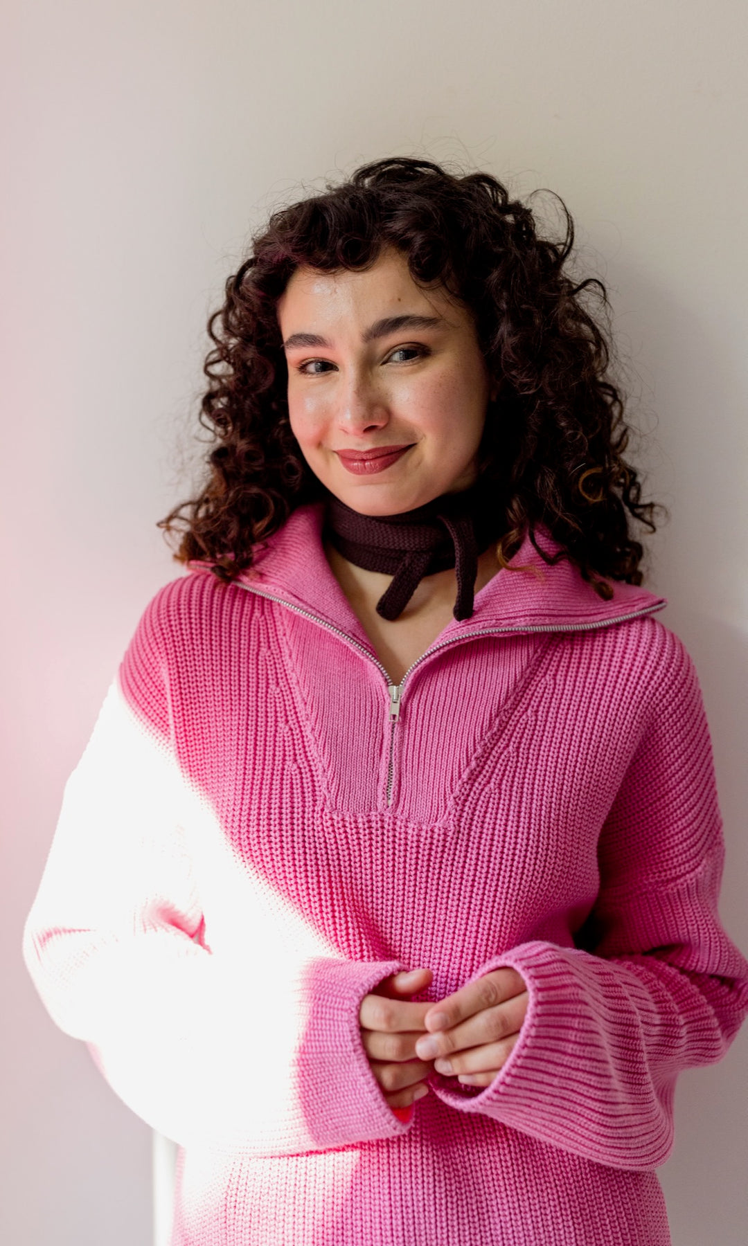 Kleiner Mini Strickschal in braun im Outfit mit Strick Pullover in pink vor heller Wand Blick gerade lächelnd