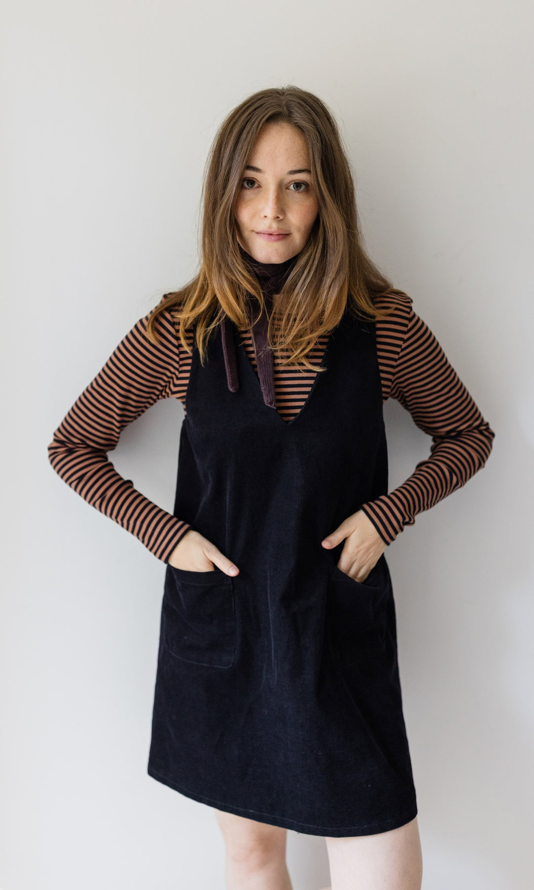 Kleiner Mini Strickschal in braun im Outfit mit braun gestreiftem Shirt, dunklem Kleid Hände in Taschen vor heller Wand Blick gerade