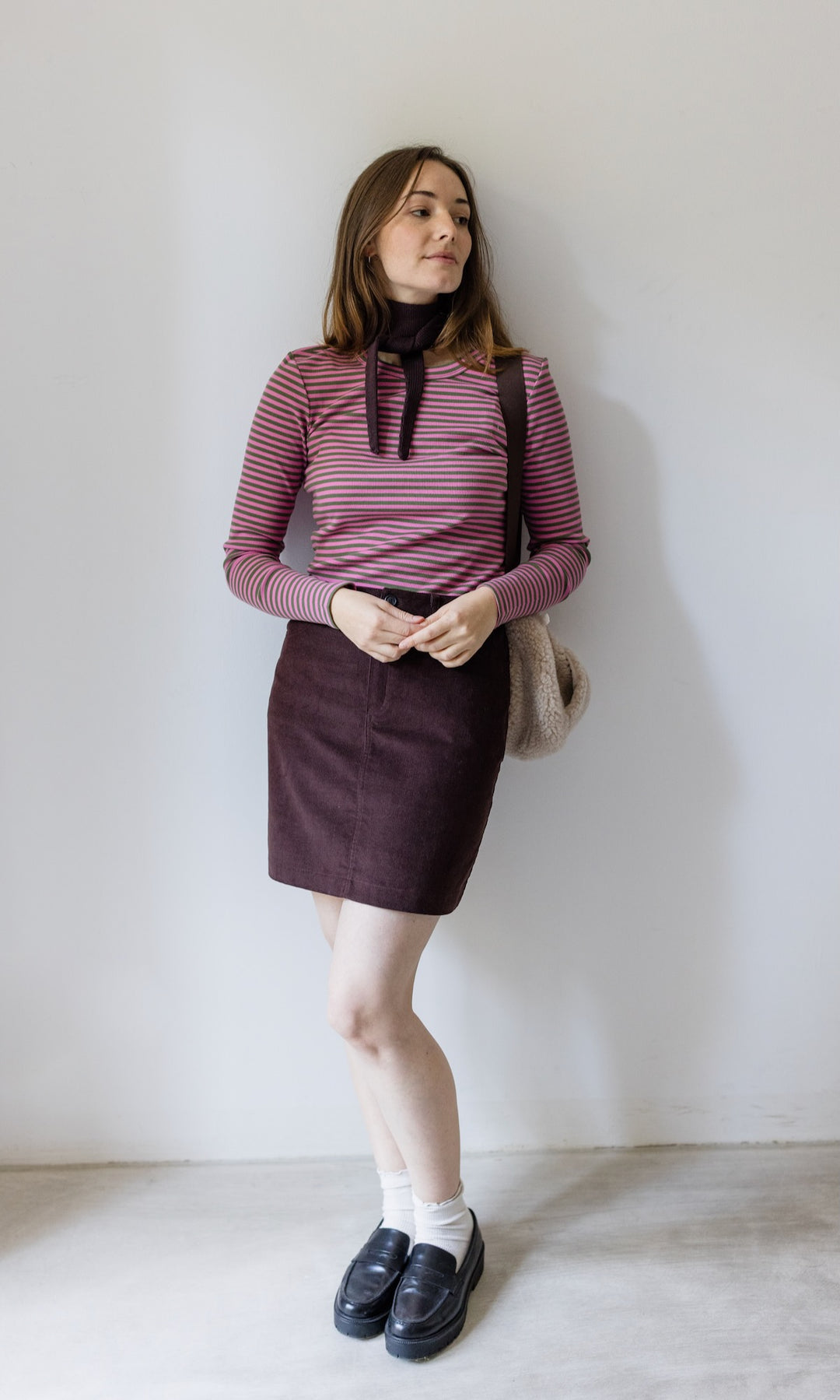 Kleiner Mini Strickschal in braun im Outfit mit rosa gestreiftem Shirt, braunem Rock, schwarzen Schuhen und Woll Handtasche angelehnt an heller Wand Blick seitlich