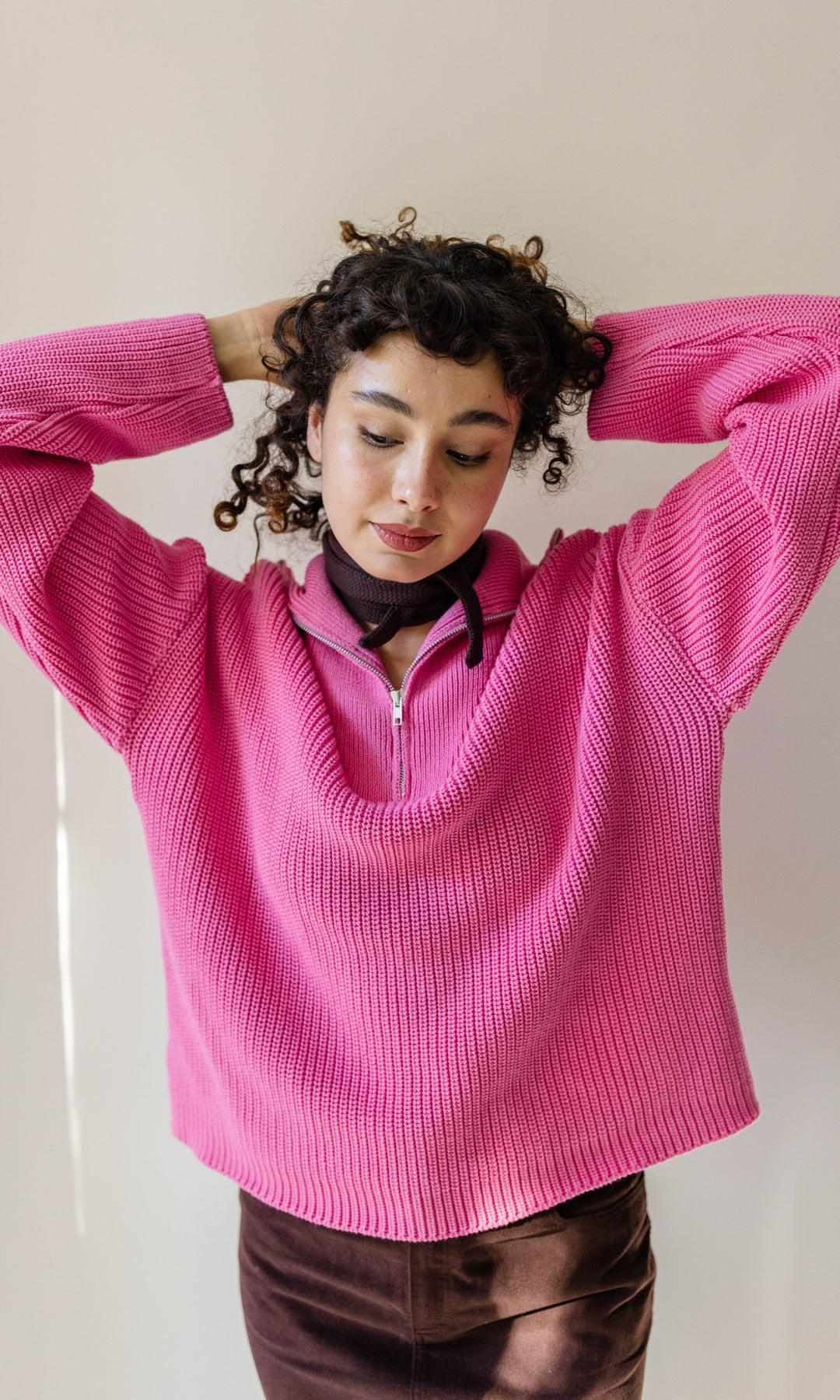 Kleiner Mini Strickschal in braun im Outfit mit Strick Pullover in pink und braunem Rock vor heller Wand Blick nach unten lächelnd