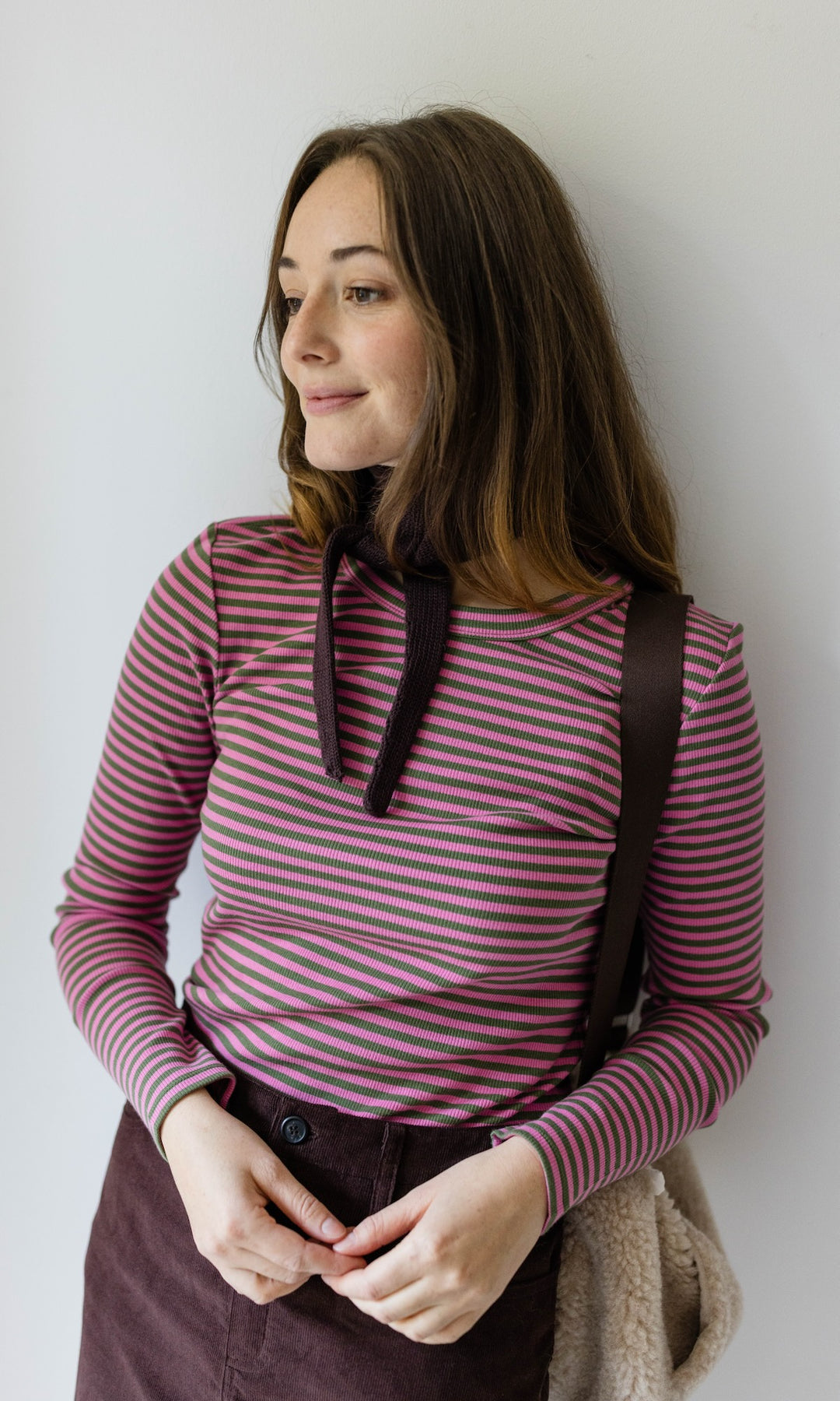 Kleiner Mini Strickschal in braun im Outfit mit rosa gestreiftem Shirt und Woll Handtasche vor heller Wand Blick seitlich