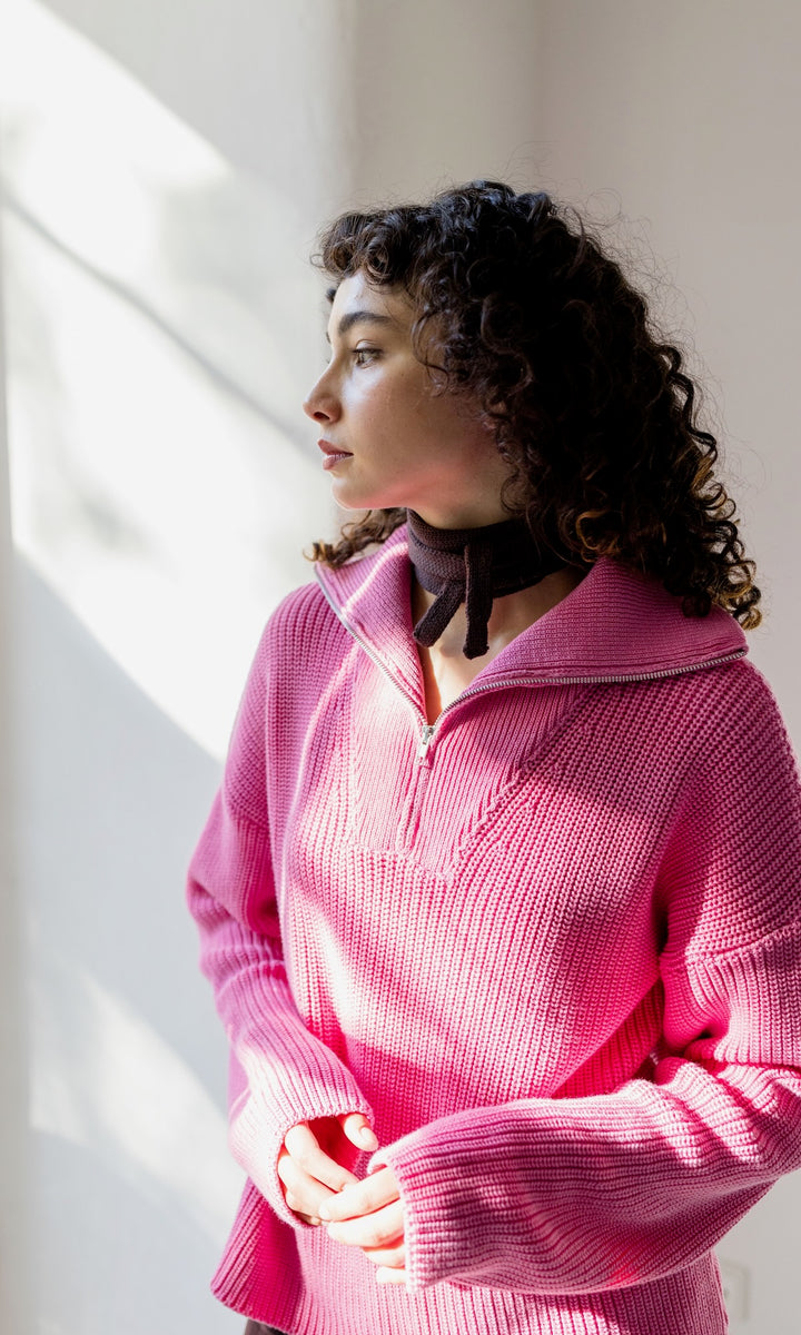 Kleiner Mini Strickschal in braun im Outfit mit Strick Pullover in pink vor heller Wand Blick seitlich zum Fenster