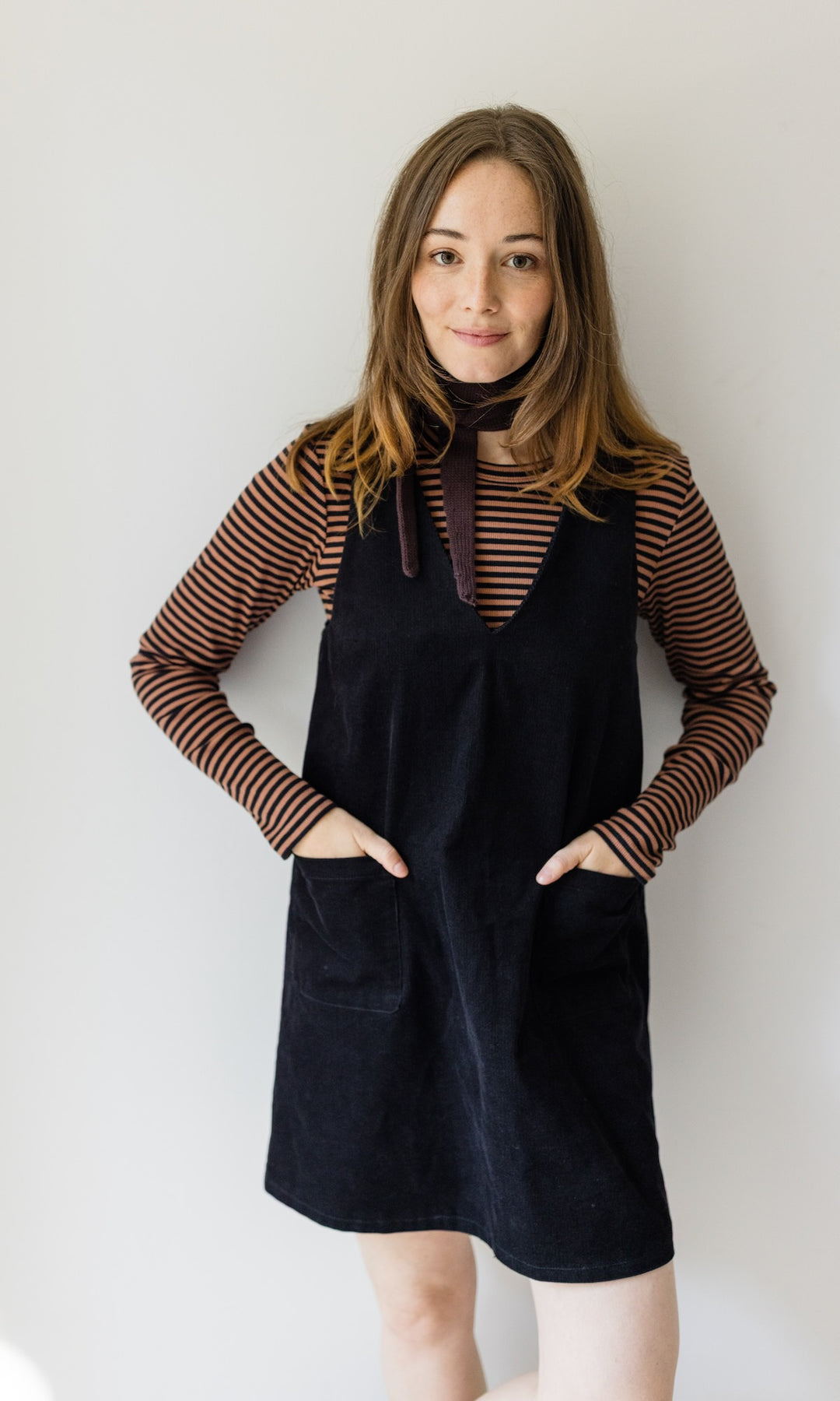 Kleiner Mini Strickschal in braun im Outfit mit braun gestreiftem Shirt, dunklem Kleid vor heller Wand Blick gerade lächelnd