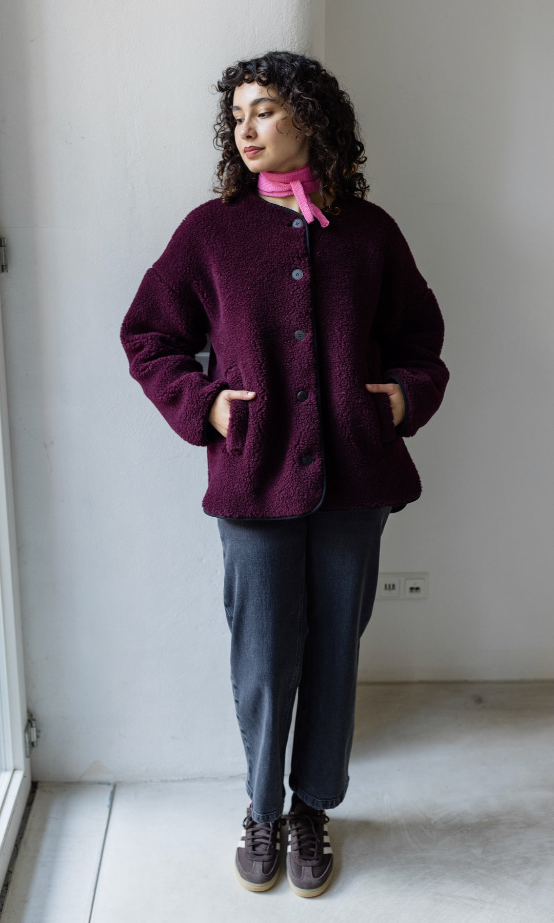 Kleiner Mini Strickschal in rosa im Outfit mit roter Schafwoll Jacke, blauer Hose und braunen Sneakers vor heller Wand Blick seitlich