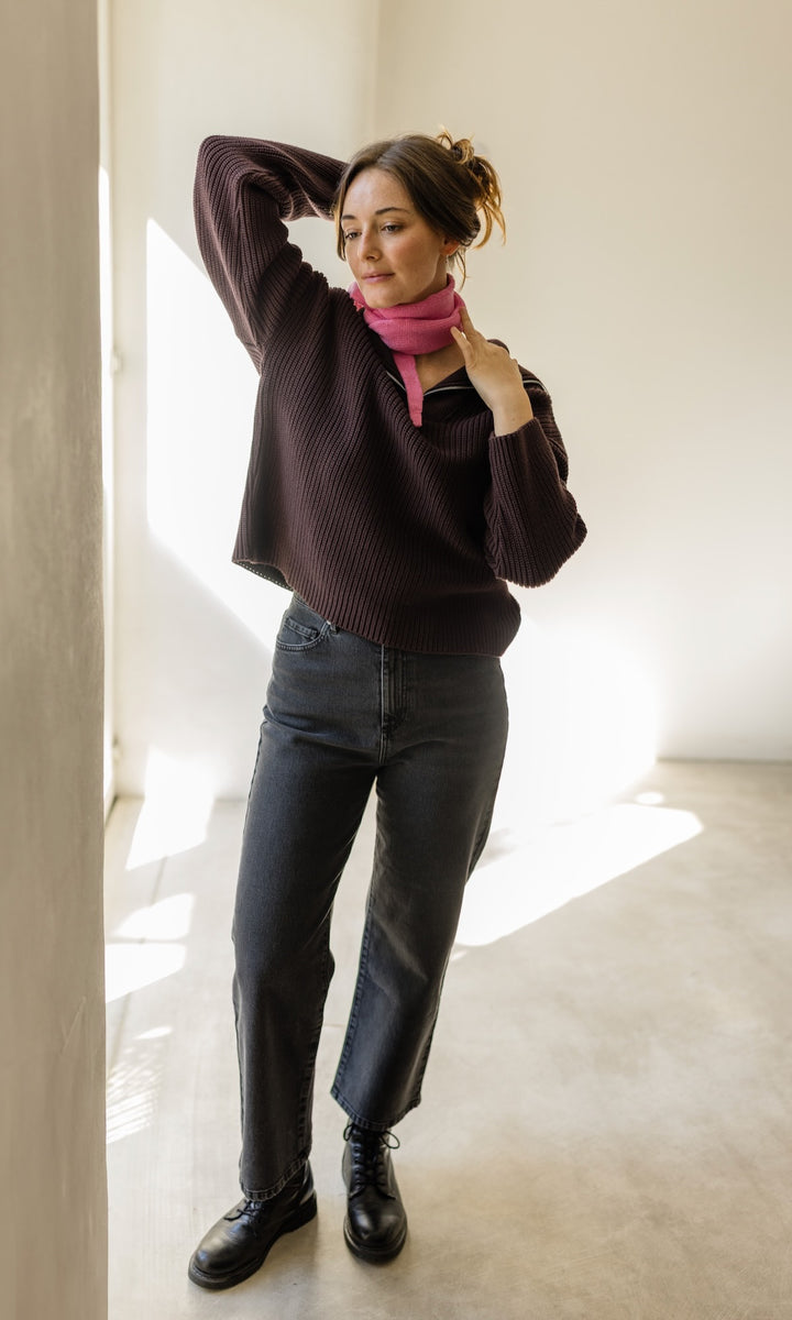 Kleiner Mini Strickschal in rosa im Outfit mit rotem Pullover, schwarzer Jeans Hose und dunklen Schuhen vor heller Wand Blick seitlich