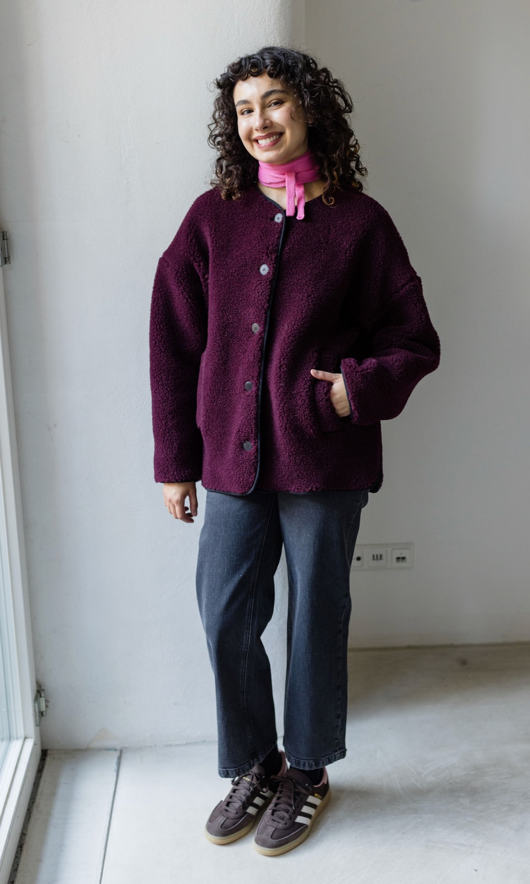 Kleiner Mini Strickschal in rosa im Outfit mit roter Schafwoll Jacke, blauer Hose und braunen Sneakers vor heller Wand Blick gerade lachend