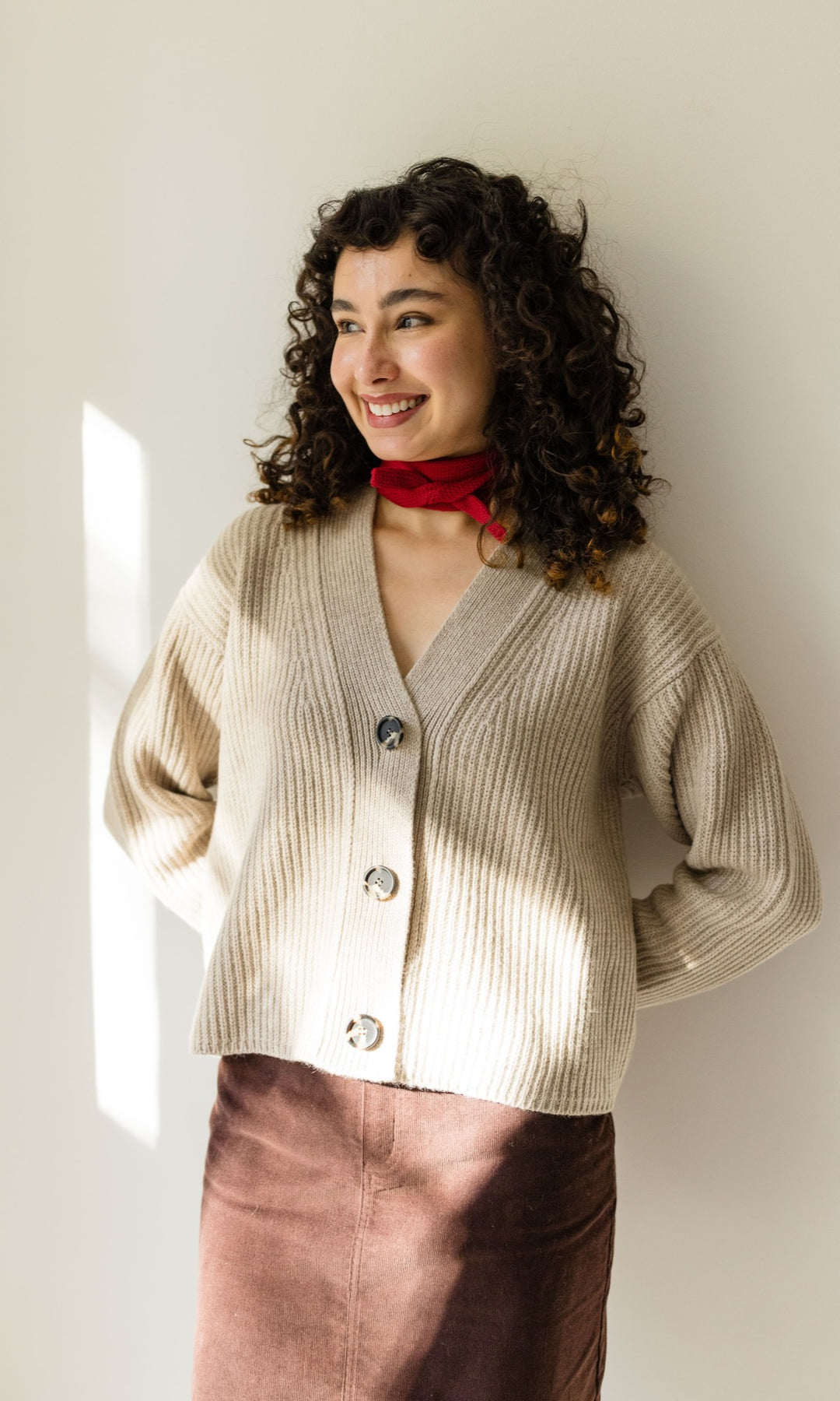 Kleiner Mini Strickschal in rot im Outfit mit hellem Strick Cardigan, braunem Rock vor heller Wand Blick seitlich lachend