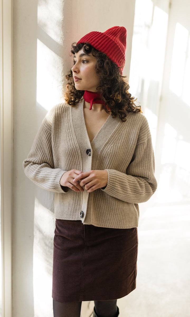 Kleiner Mini Strickschal in rot im Outfit mit hellem Strick Cardigan und roter Strick Mütze, braunem Rock und dunkler Strumpfhose vor heller Wand Blick seitlich