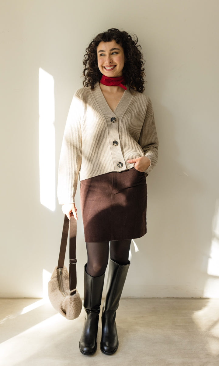 Kleiner Mini Strickschal in rot im Outfit mit hellem Strick Cardigan, braunem Rock und dunkler Strumpfhose, schwarzen Stiefeln und Woll Handtasche vor heller Wand Blick seitlich lachend