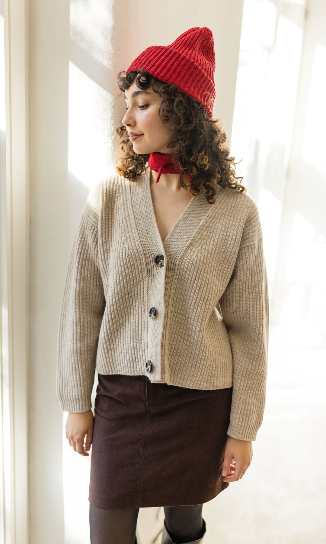 Kleiner Mini Strickschal in rot im Outfit mit hellem Strick Cardigan und roter Strickmütze, braunem Rock und dunkler Strumpfhose vor heller Wand Blick seitlich