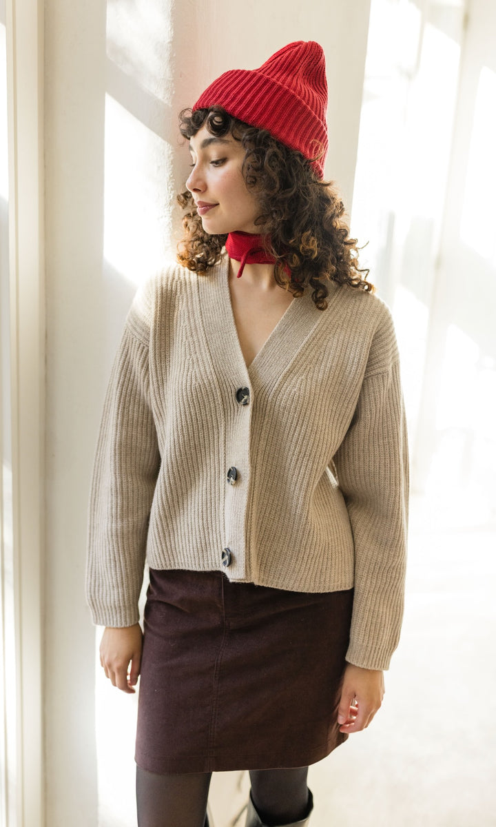 Kleiner Mini Strickschal in rot im Outfit mit hellem Strick Cardigan und roter Strickmütze, braunem Rock und dunkler Strumpfhose vor heller Wand Blick seitlich