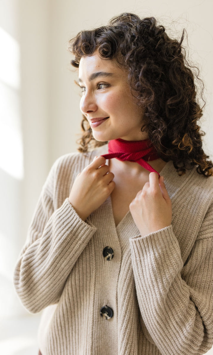 Kleiner Mini Strickschal in rot im Outfit mit hellem Strick Cardigan vor heller Wand Blick seitlich lächelnd