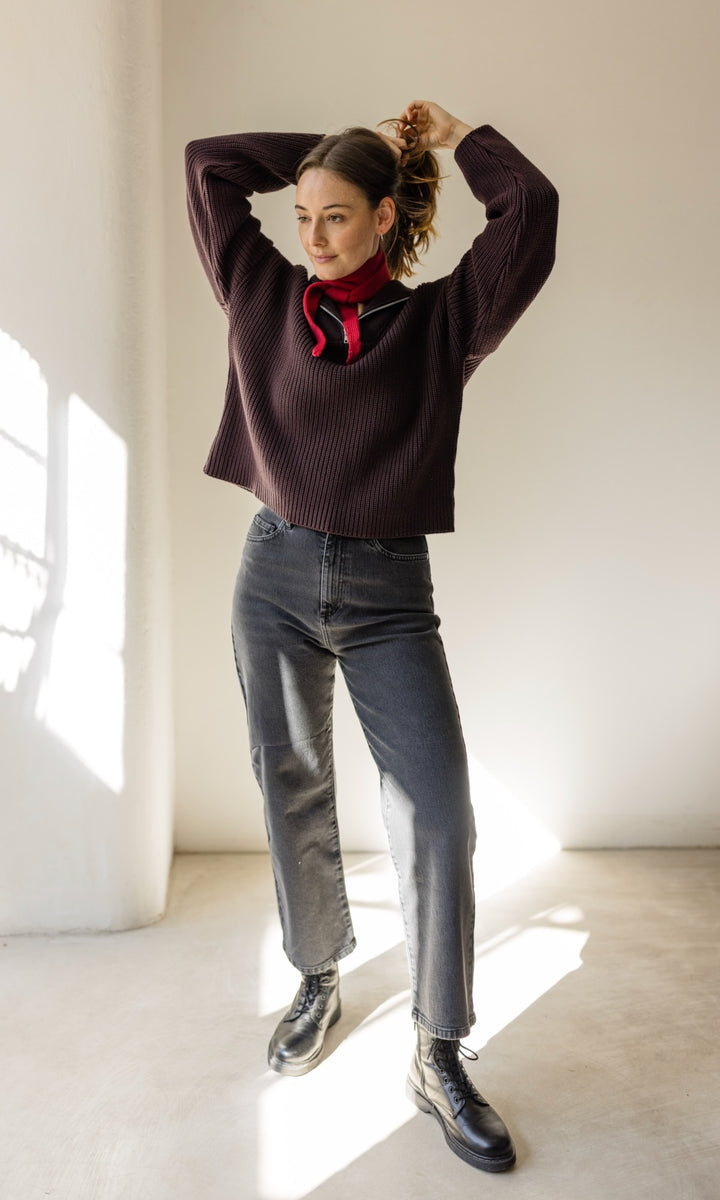 Kleiner Mini Strickschal in rot im Outfit mit rotem Pullover, schwarzer Jeans Hose und dunklen Stiefeln vor heller Wand Blick seitlich lächelnd