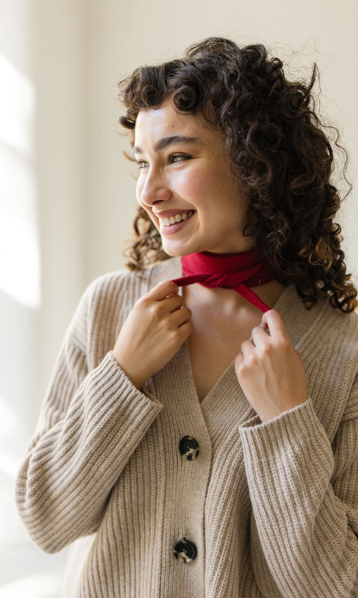 Kleiner Mini Strickschal in rot im Outfit mit hellem Strick Cardigan vor heller Wand Blick seitlich lachend