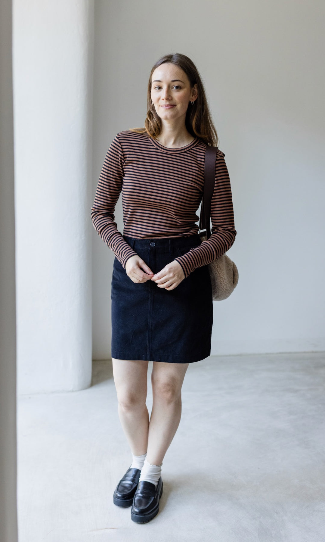 Langarm Stillshirt Zoe in braun gestreift kombiniert mit schwarzem Rock und Tasche aus Schafwolle vor grauer Wand von vorne Blick gerade lächelnd