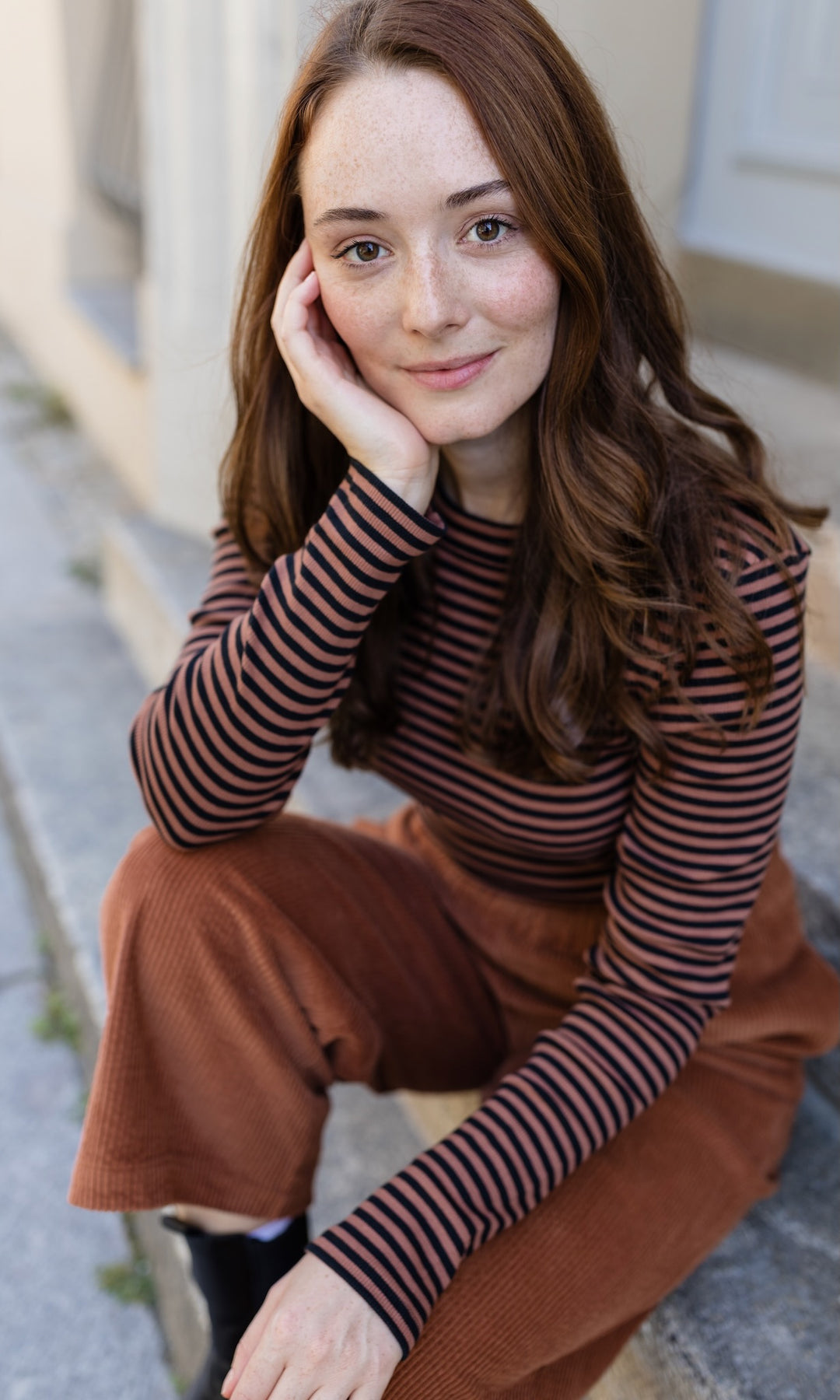 Langarm Stillshirt Zoe in braun gestreift kombiniert mit brauner Hose sitzend vor grauer Wand im Herbst seitlich Blick gerade