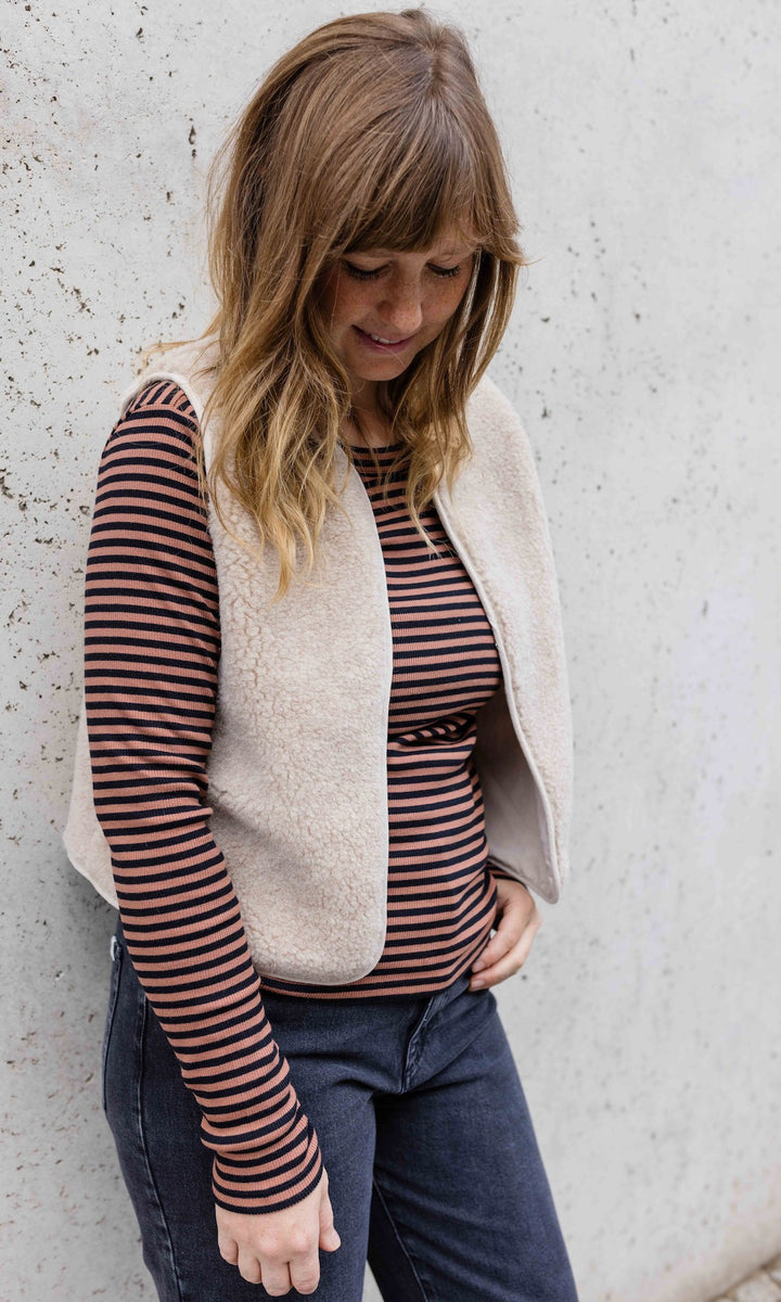 Langarm Stillshirt Zoe in braun gestreift im Winter kombiniert mit blauer Jeans und heller Schafwoll-Weste vor grauer Wand seitlich Blick nach unten lächelnd