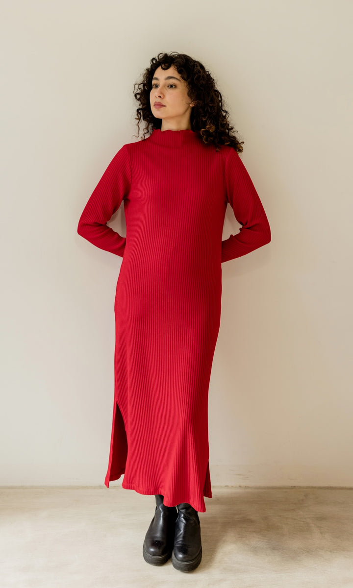 Langes Strickkleid Jil in rot mit schwarzen Stiefeln angelehnt an heller Wand Blick seitlich