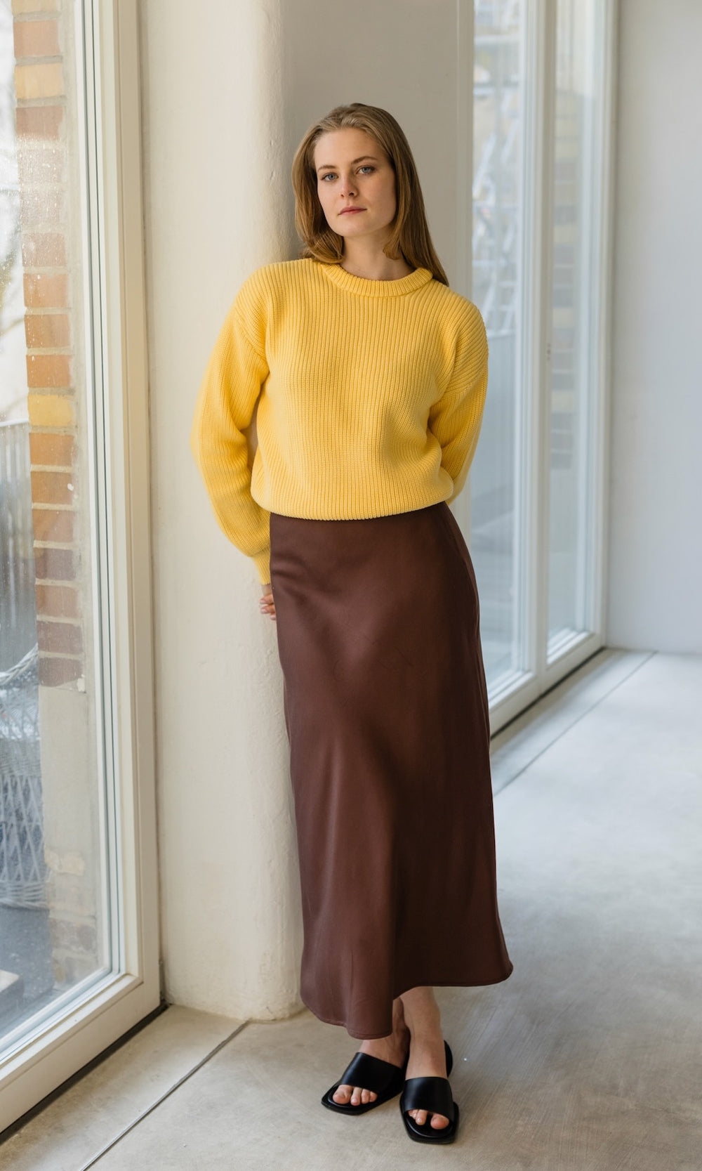 Senfgelber Strickpullover aus Bio-Baumwolle kombiniert mit langem braunen Rock und schwarzen Sandalen.