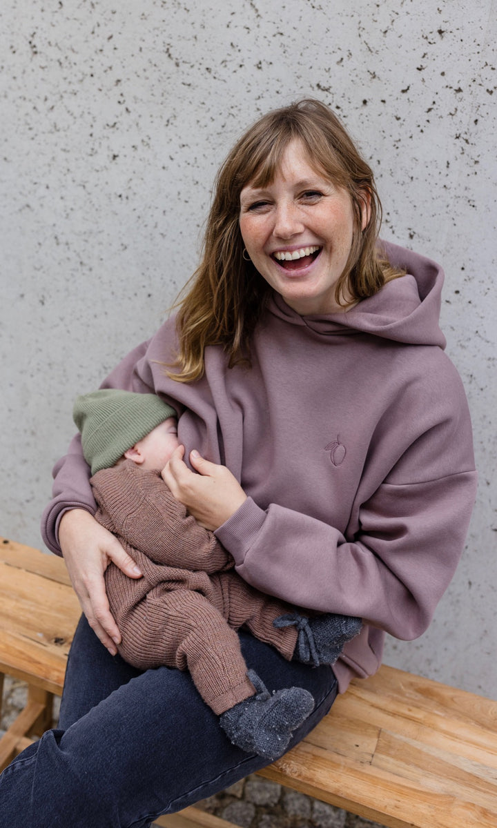 Still Hoodie in aubergine rot sitzend auf Bank mit Baby beim Stillen vor grauer Wand lachend