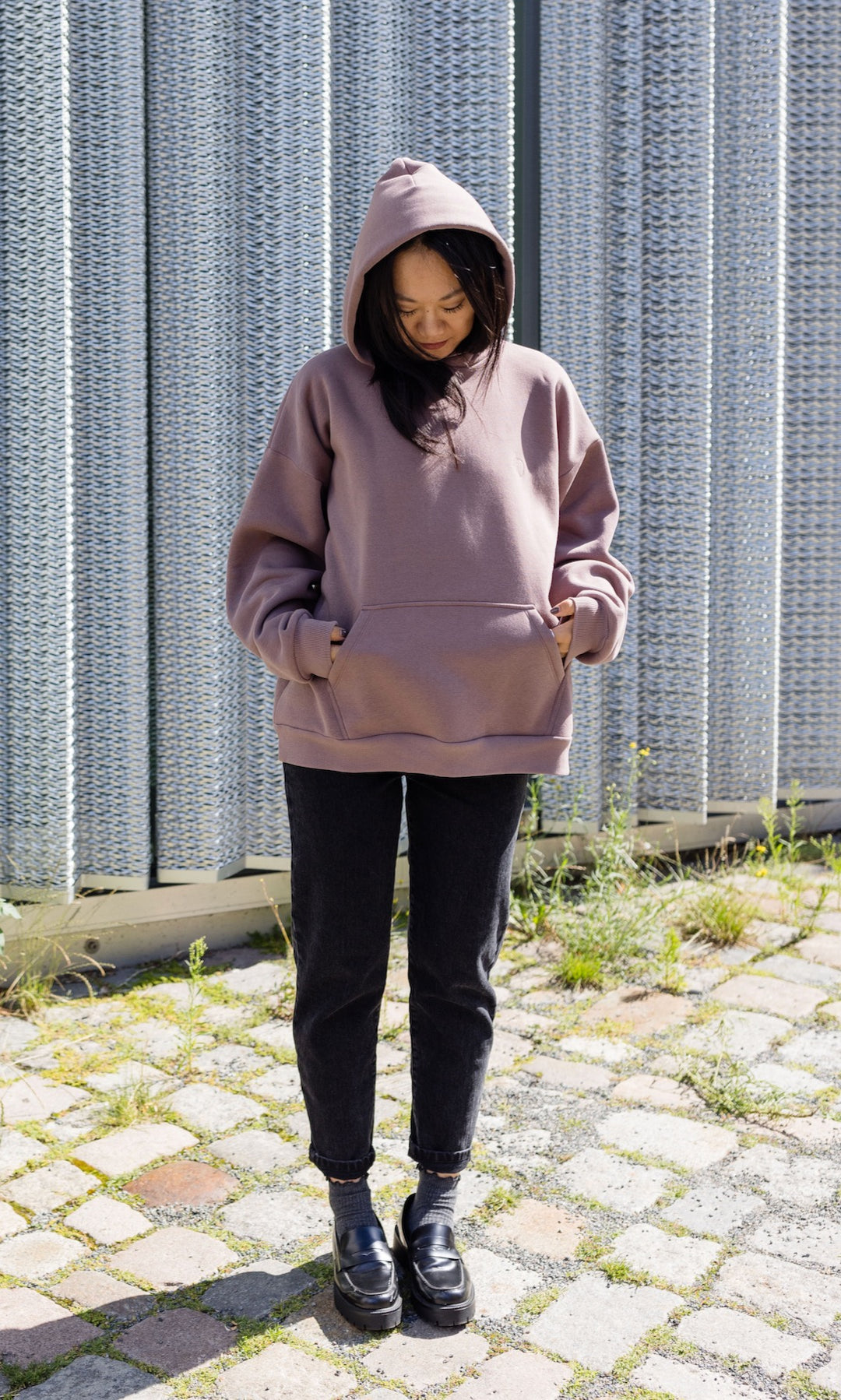 Still Hoodie in aubergine rot stehend mit schwarzer Hose und Stiefeln vor grauer Wand Kapuze auf Kopf Hände in Tasche von vorne Blick nach unten 