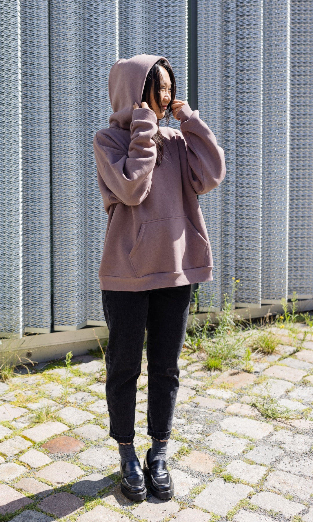 Still Hoodie in aubergine rot stehend mit schwarzer Hose und Stiefeln vor grauer Wand Kapuze auf Kopf von vorne Blick seitlich 