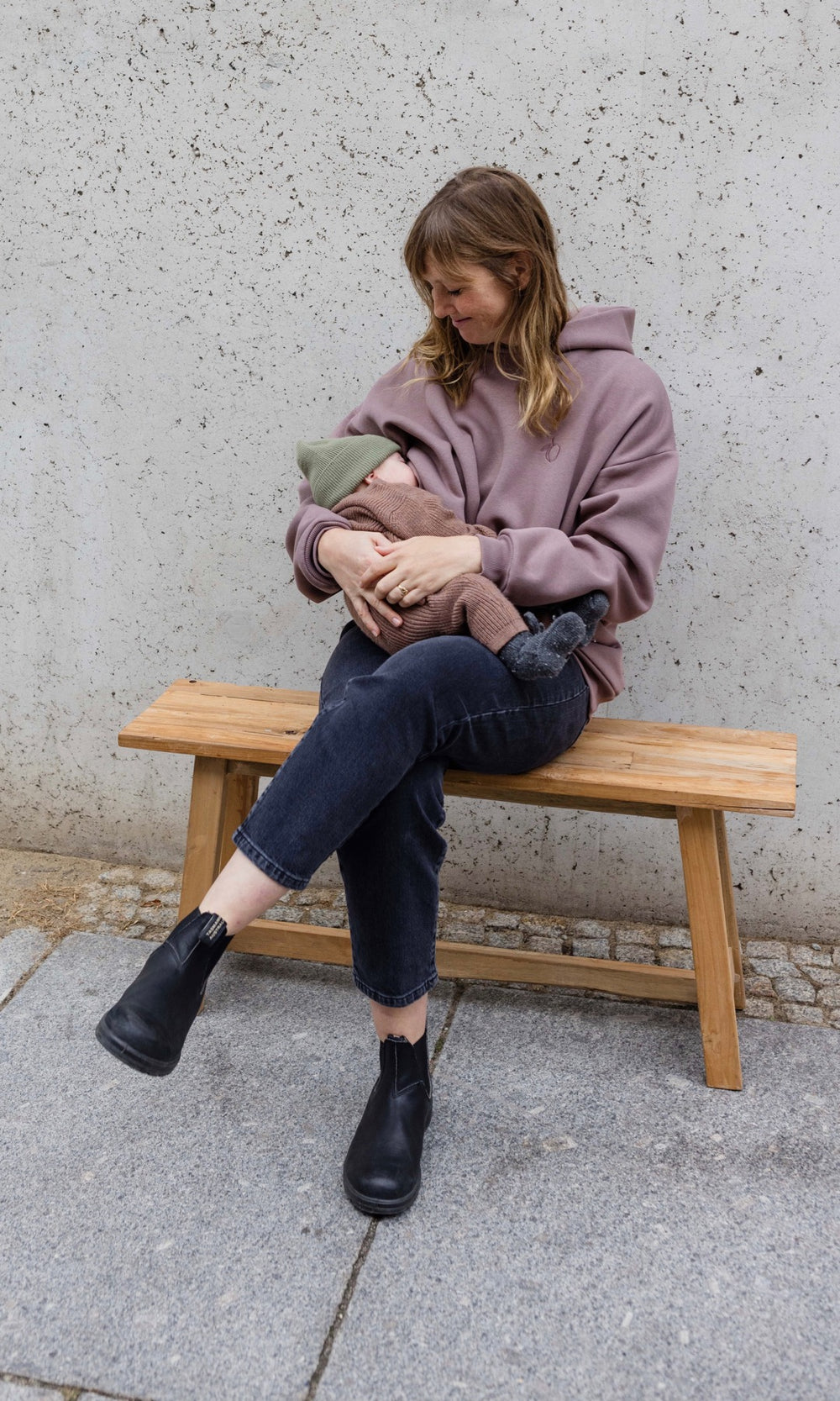 Still Hoodie in aubergine rot sitzend auf Bank mit Baby beim Stillen vor grauer Wand von vorne mit Blick auf Kind