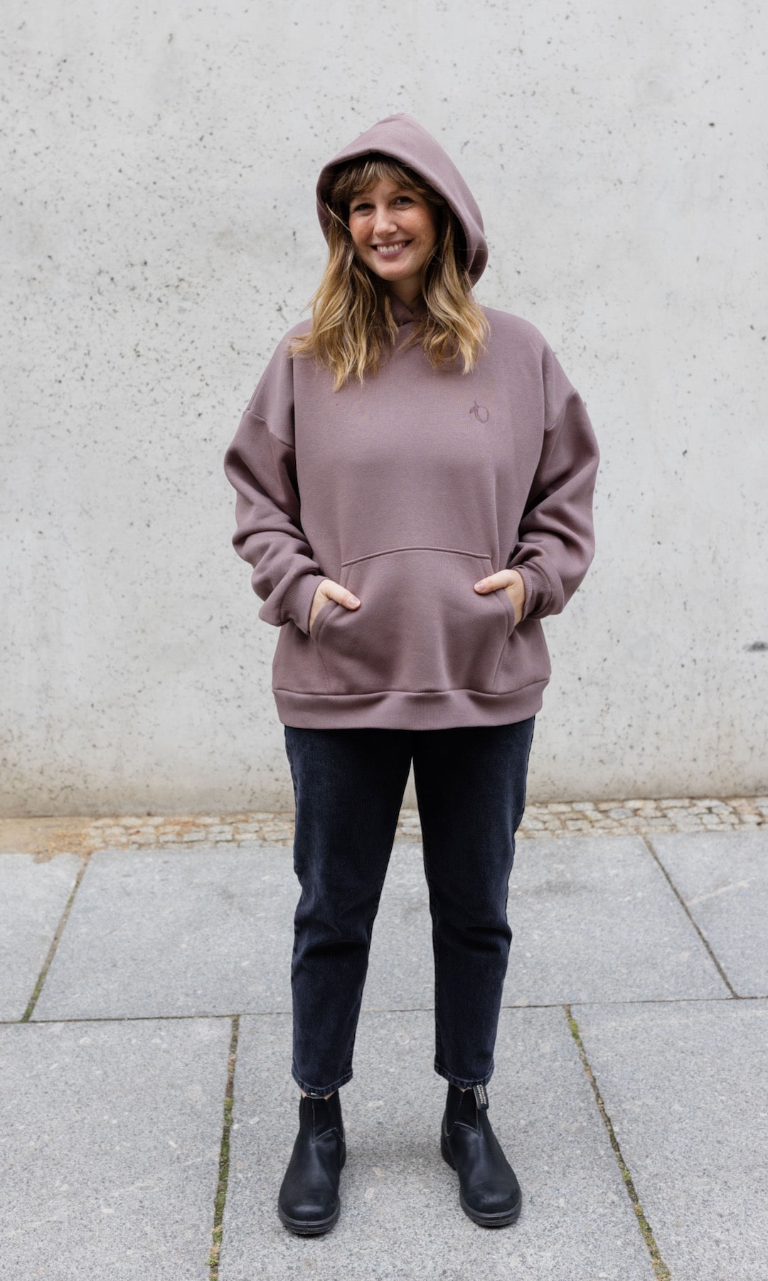 Still Hoodie in aubergine rot stehend mit schwarzer Hose und Stiefeln vor grauer Wand Kapuze auf Kopf Hände in Tasche von vorne lächelnd