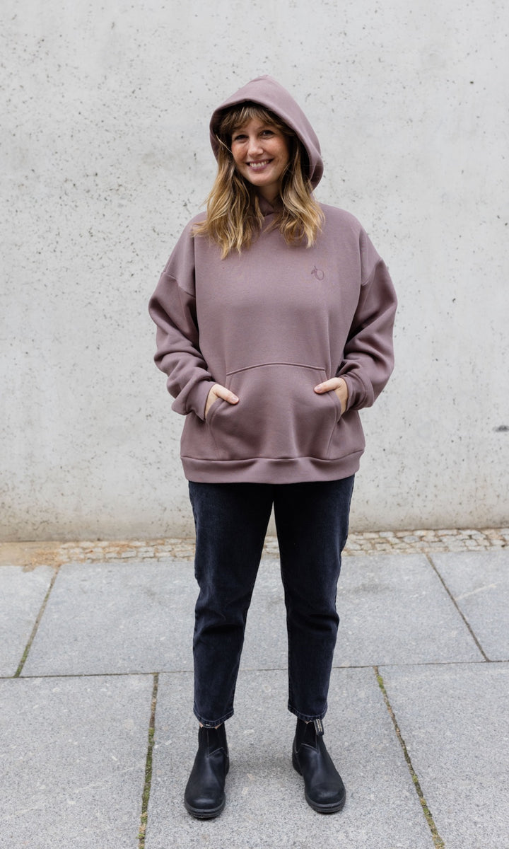 Still Hoodie in aubergine rot stehend mit schwarzer Hose und Stiefeln vor grauer Wand Kapuze auf Kopf Hände in Tasche von vorne lächelnd