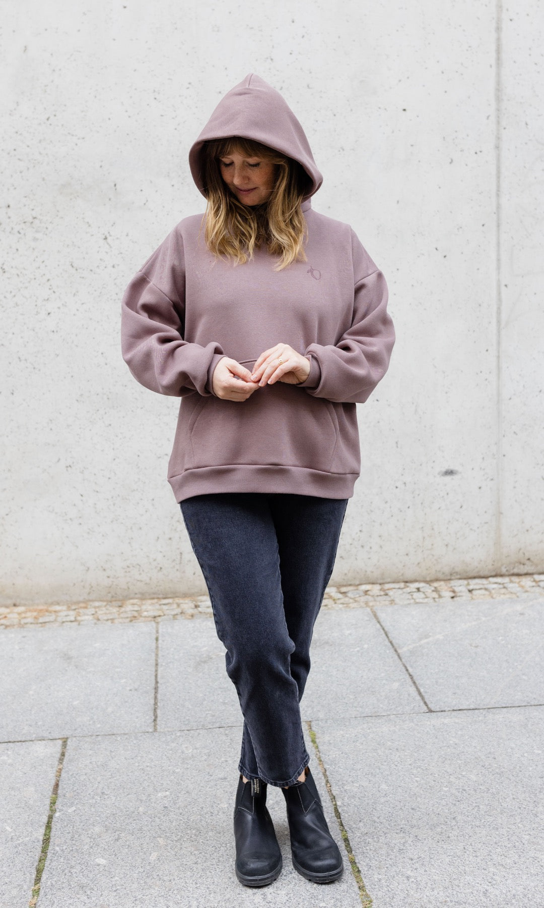 Still Hoodie in aubergine rot stehend mit schwarzer Hose und Stiefeln vor grauer Wand Kapuze auf Kopf Blick nach unten