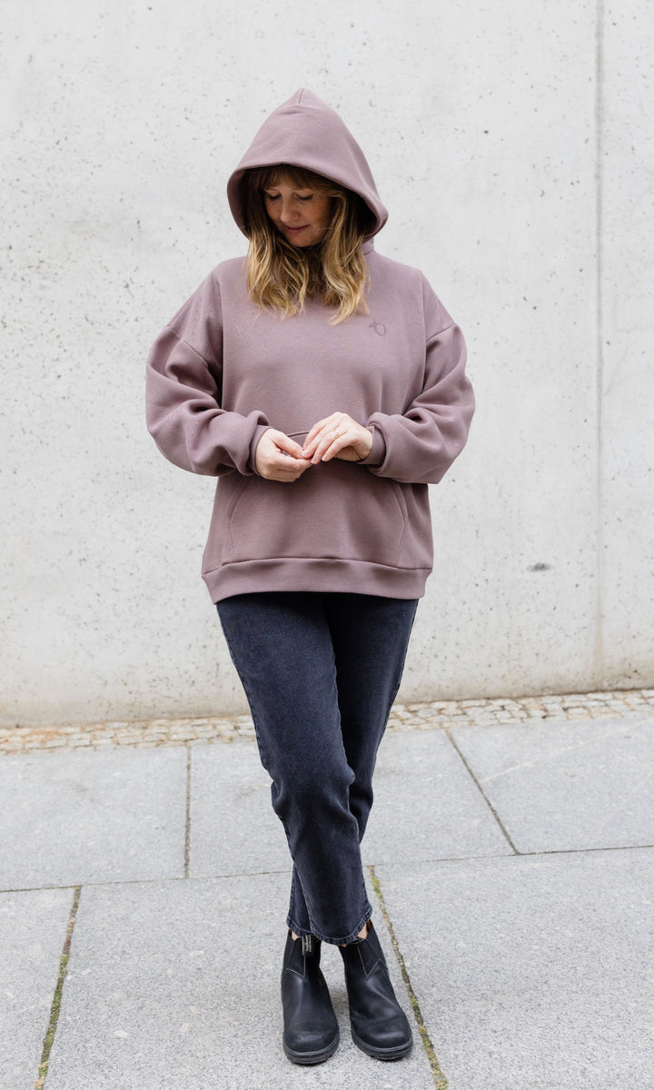 Still Hoodie in aubergine rot stehend mit schwarzer Hose und Stiefeln vor grauer Wand Kapuze auf Kopf Blick nach unten