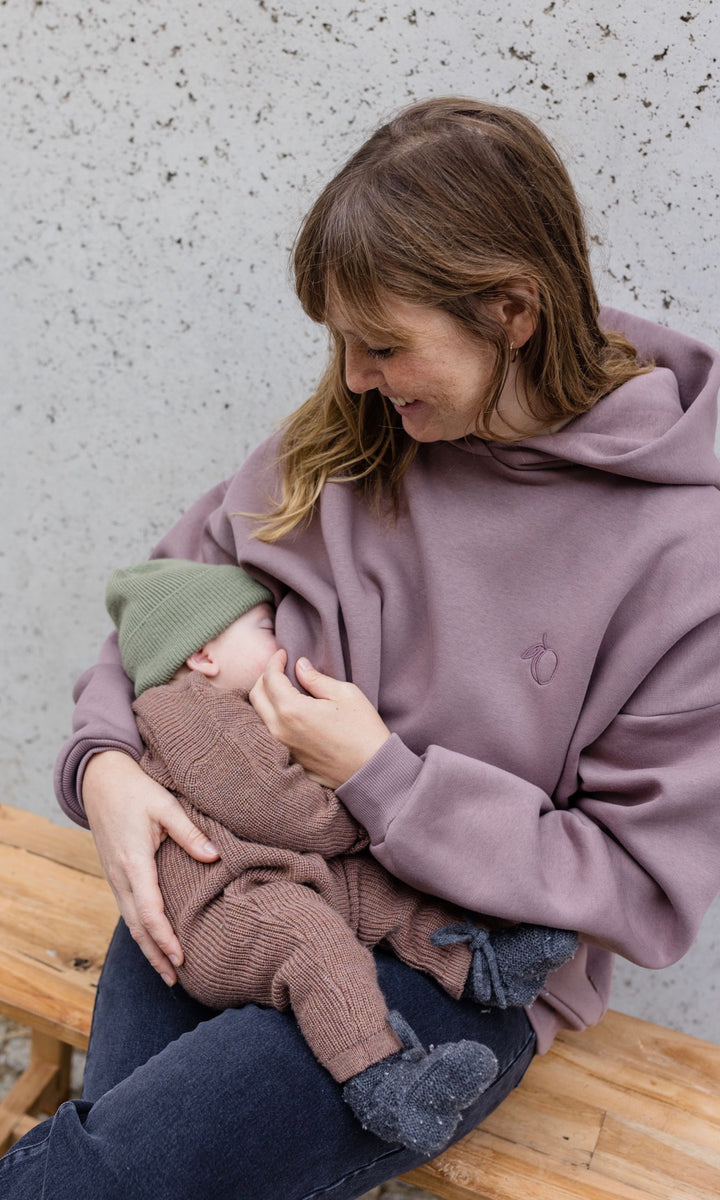 Still Hoodie in aubergine rot sitzend auf Bank mit Baby beim Stillen vor grauer Wand Blick auf Kind