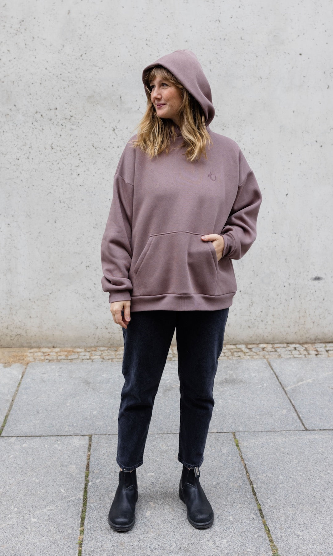 Still Hoodie in aubergine rot stehend mit schwarzer Hose und Stiefeln vor grauer Wand Kapuze auf Kopf Hand in Tasche von vorne Blick seitlich 