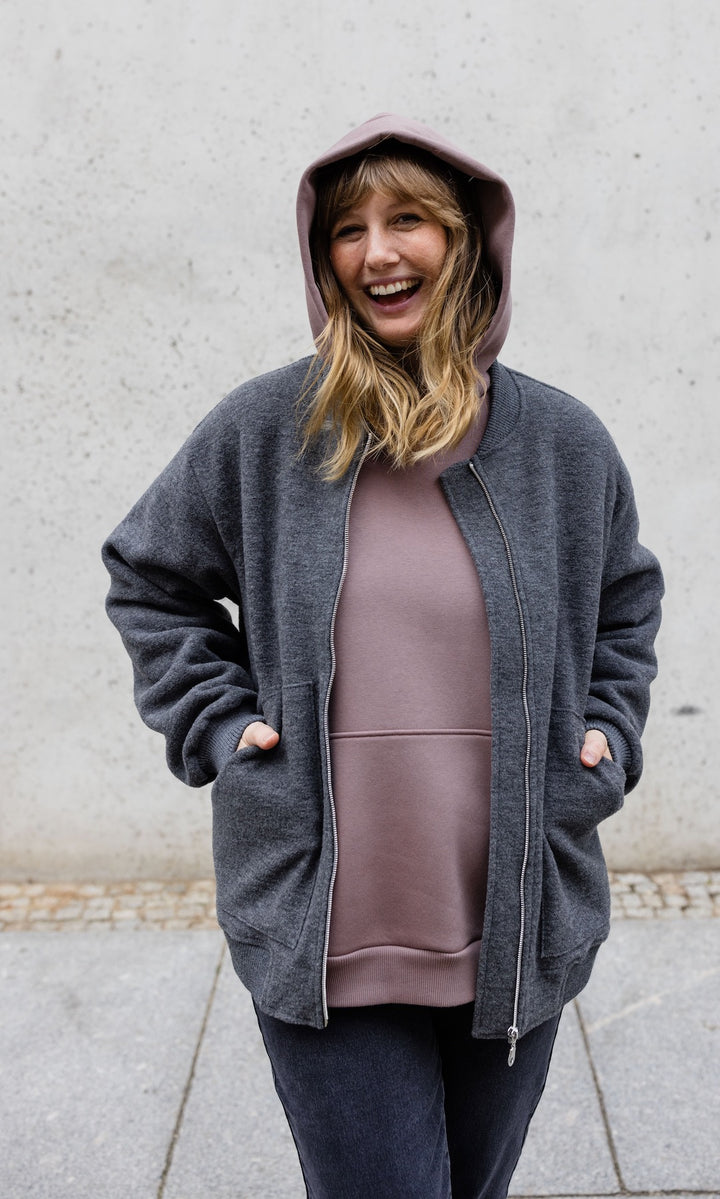 Still Hoodie in aubergine rot stehend im Herbst mit schwarzer Hose und grauer Jacke vor grauer Wand Kapuze auf Kopf Hände in Tasche der Jacke von vorne lachend